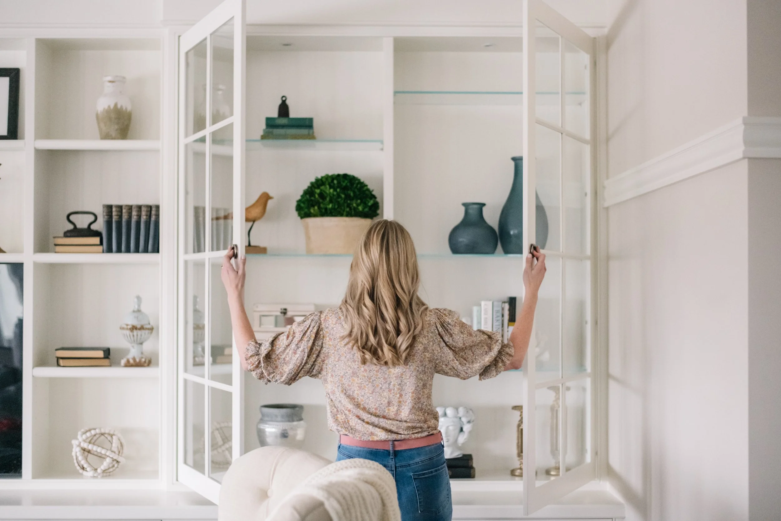 A woman opening glass doors on a white cabinet filled with decorative items and books in a living room.