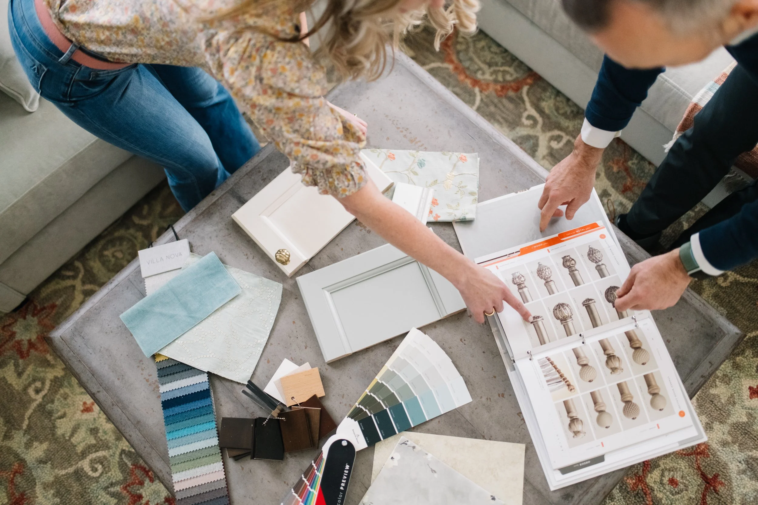 Two people examining paint color samples, cabinet door samples, and fabric swatches on a coffee table in a home interior design setting.