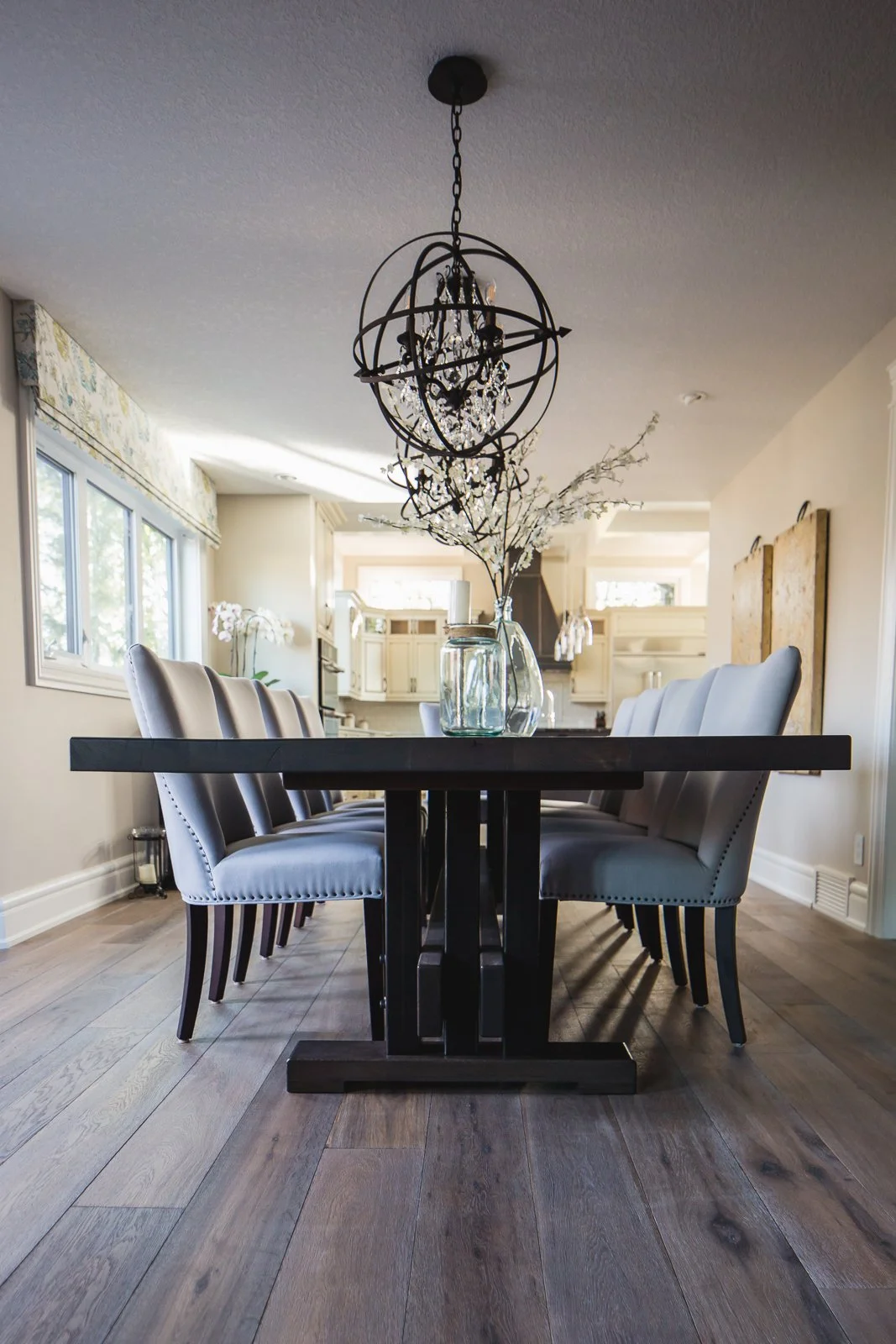 Empty dining room with a rectangular black table, white upholstered chairs, and a chandelier hanging above, in a bright open space.