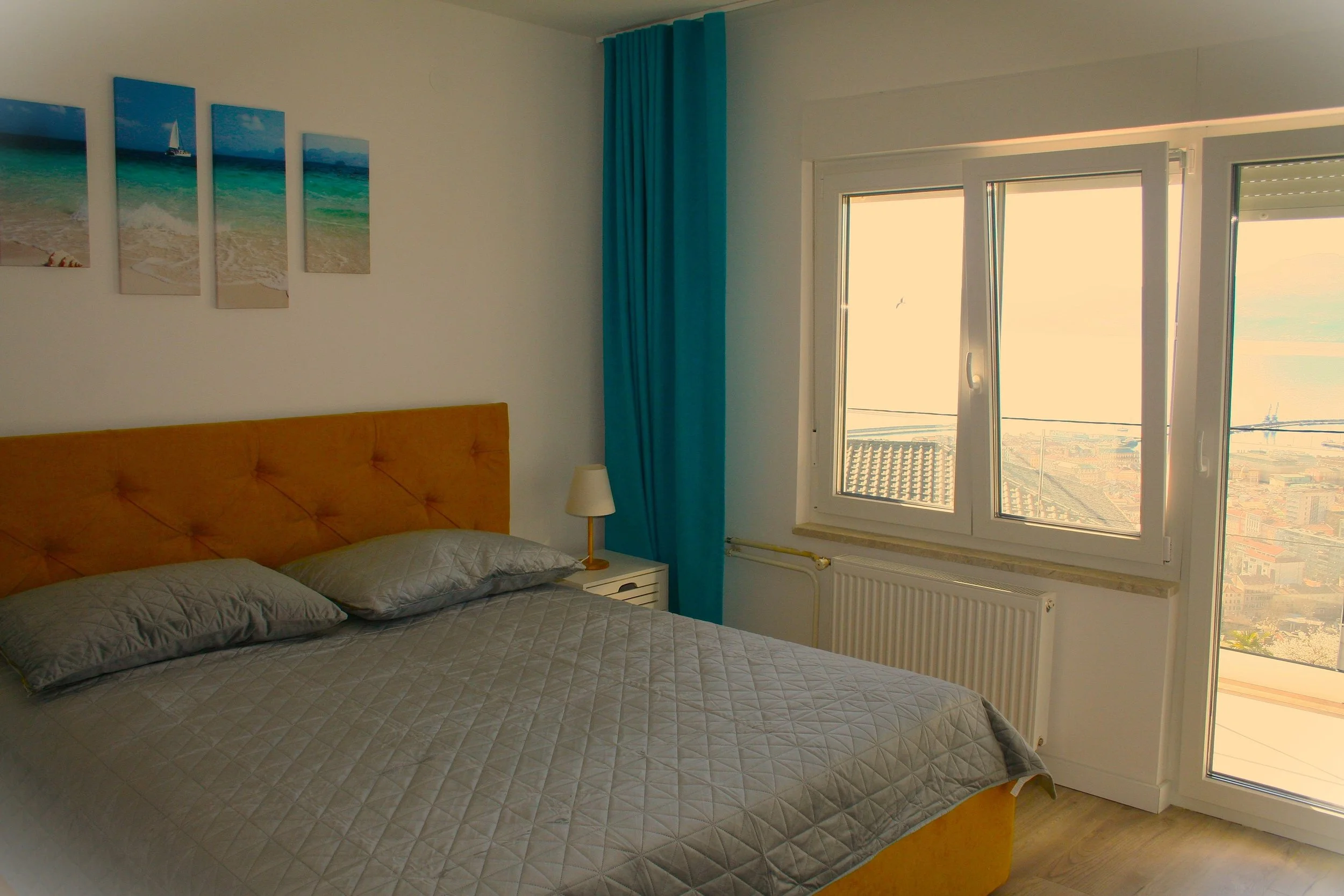 A bedroom with a bed, gray quilt, gray pillows, a small white nightstand with a lamp, blue curtains, and an open window showing an outdoor view of a cityscape and sky.