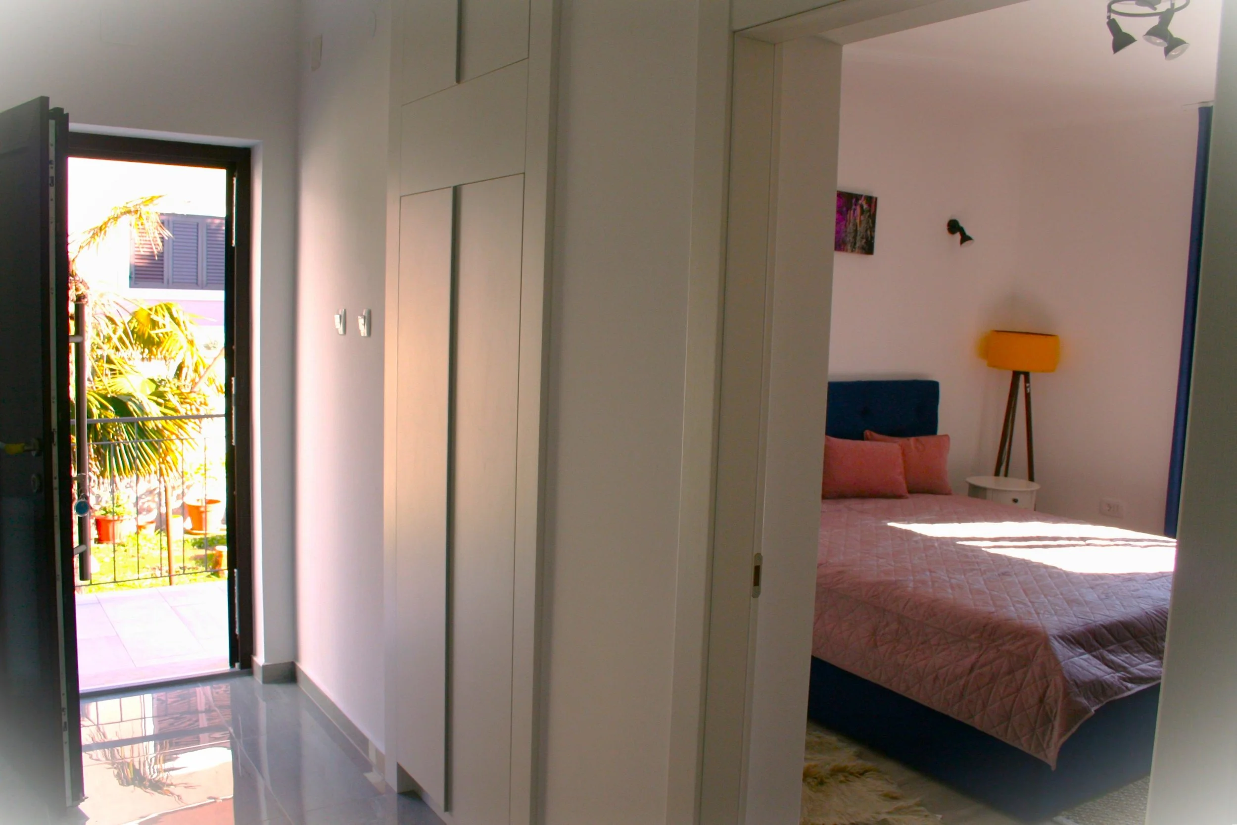 Interior view of a bedroom with a bed, pink bedding, orange pillows, a white bedside table, a yellow floor lamp, and a window with sunlight; part of a hallway with white cabinets and an open door leading to a balcony with greenery and a building outside.