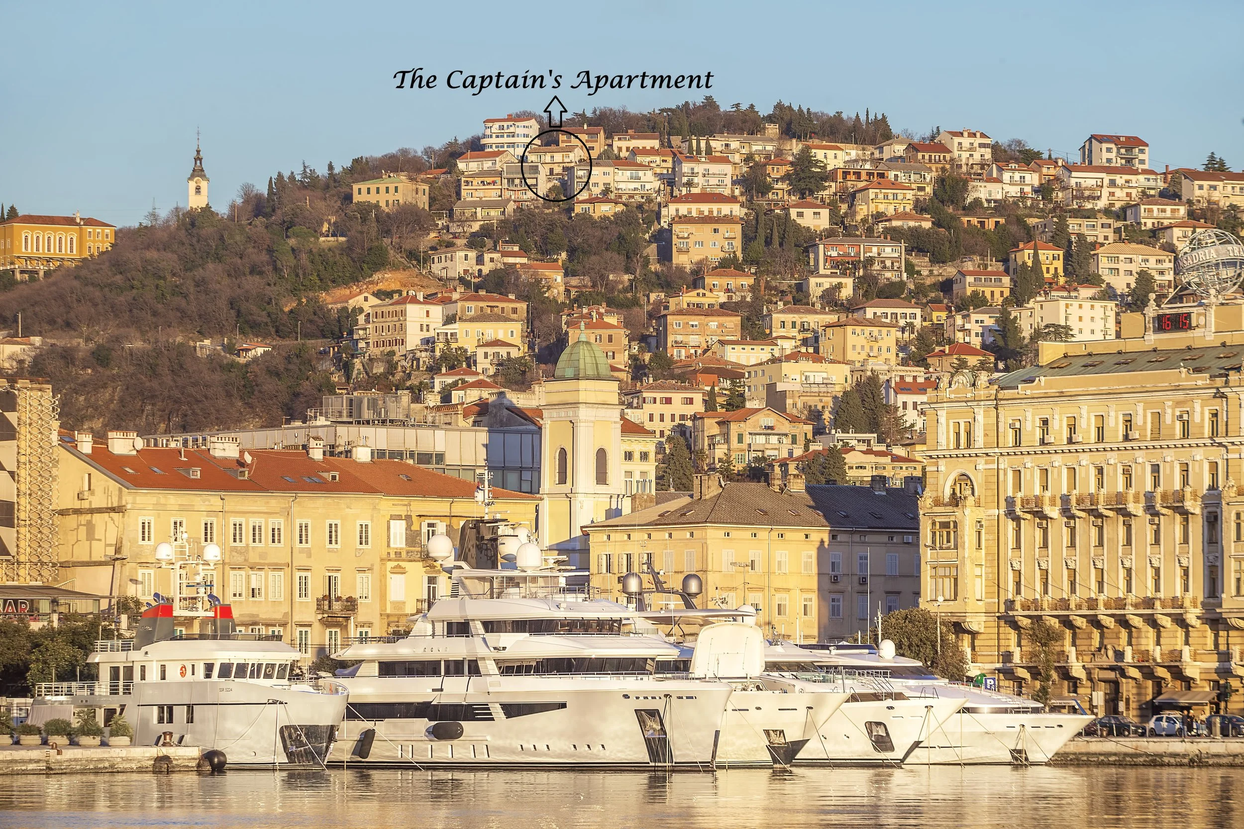 View of a harbor with luxury yachts docked in front of historic buildings, hillside with numerous colorful houses, and a church steeple in the background, labeled 'The Captain's Apartment' at the top of the hill.