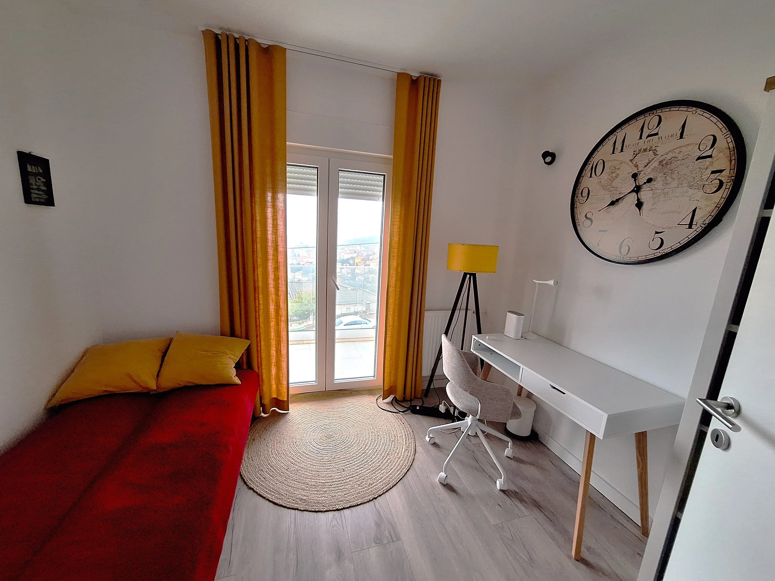 A living room corner with a red sofa, yellow cushions, yellow curtains, a glass door leading to a balcony, a round rug, a white desk, a gray office chair, a yellow floor lamp, a small white speaker, and a large wall clock with a world map design.