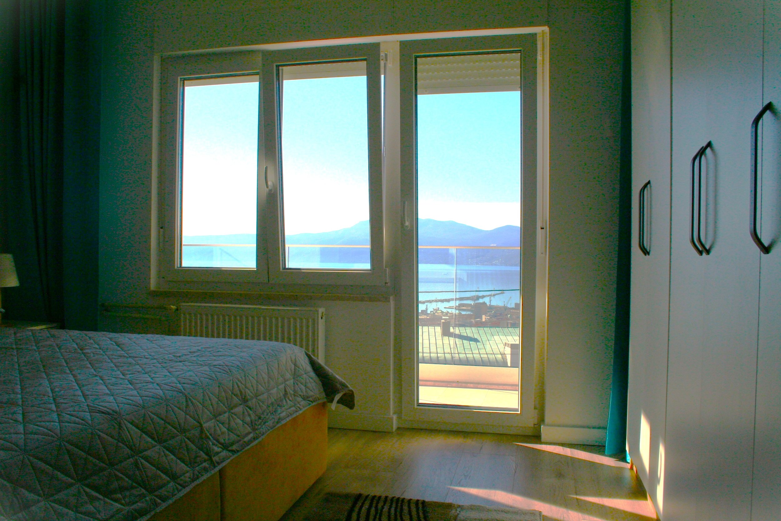 Bedroom with large window and balcony door showing a scenic view of mountains and water outside, bed with a quilted cover, and a white wardrobe.