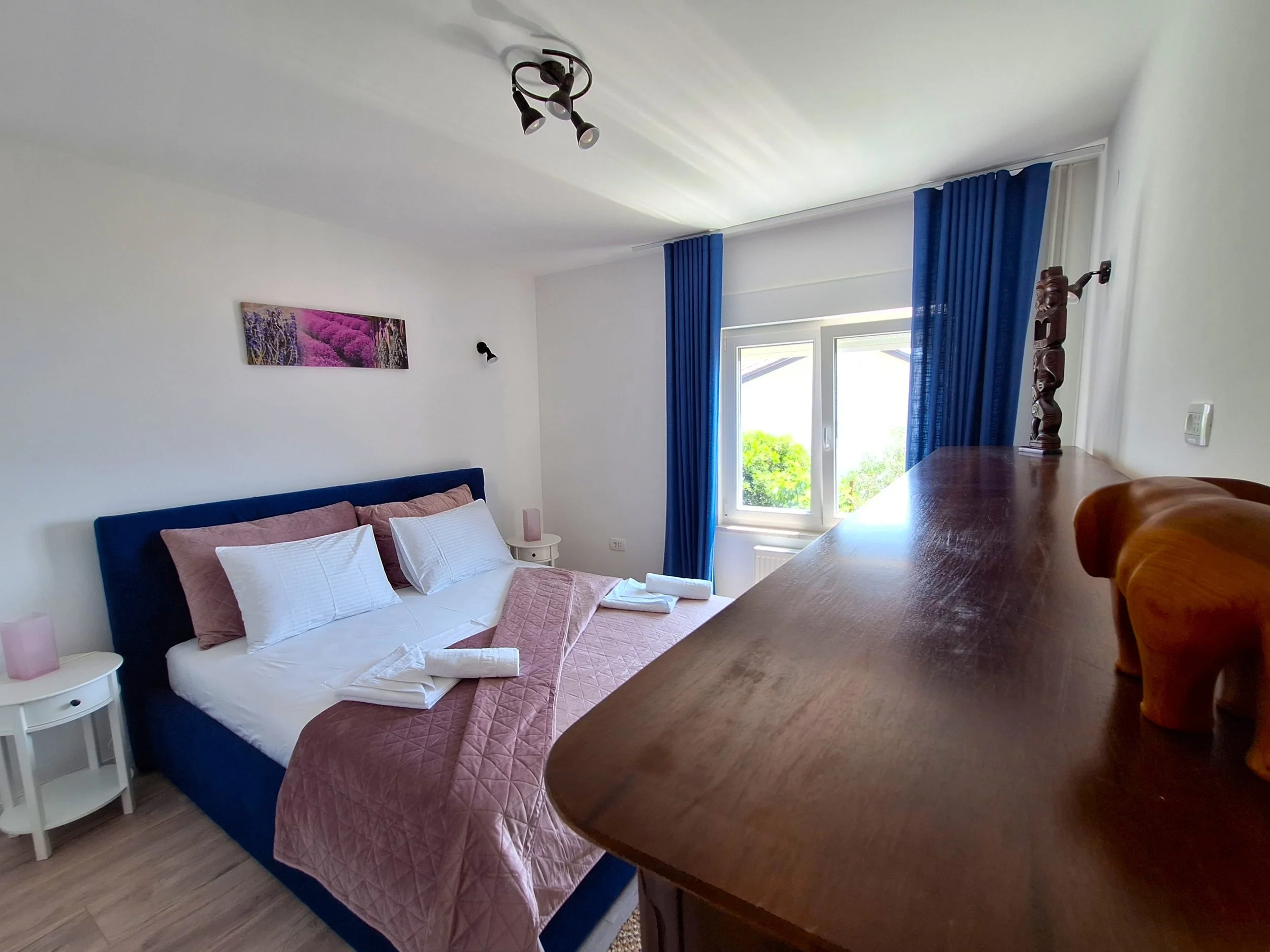 A bedroom with a bed, pink quilt, white pillows, and a blue headboard. There are blue curtains, a window showing green trees outside, and wall decorations including a mounted lamp and artwork. A wooden dresser with a carved elephant figurine is in the foreground.