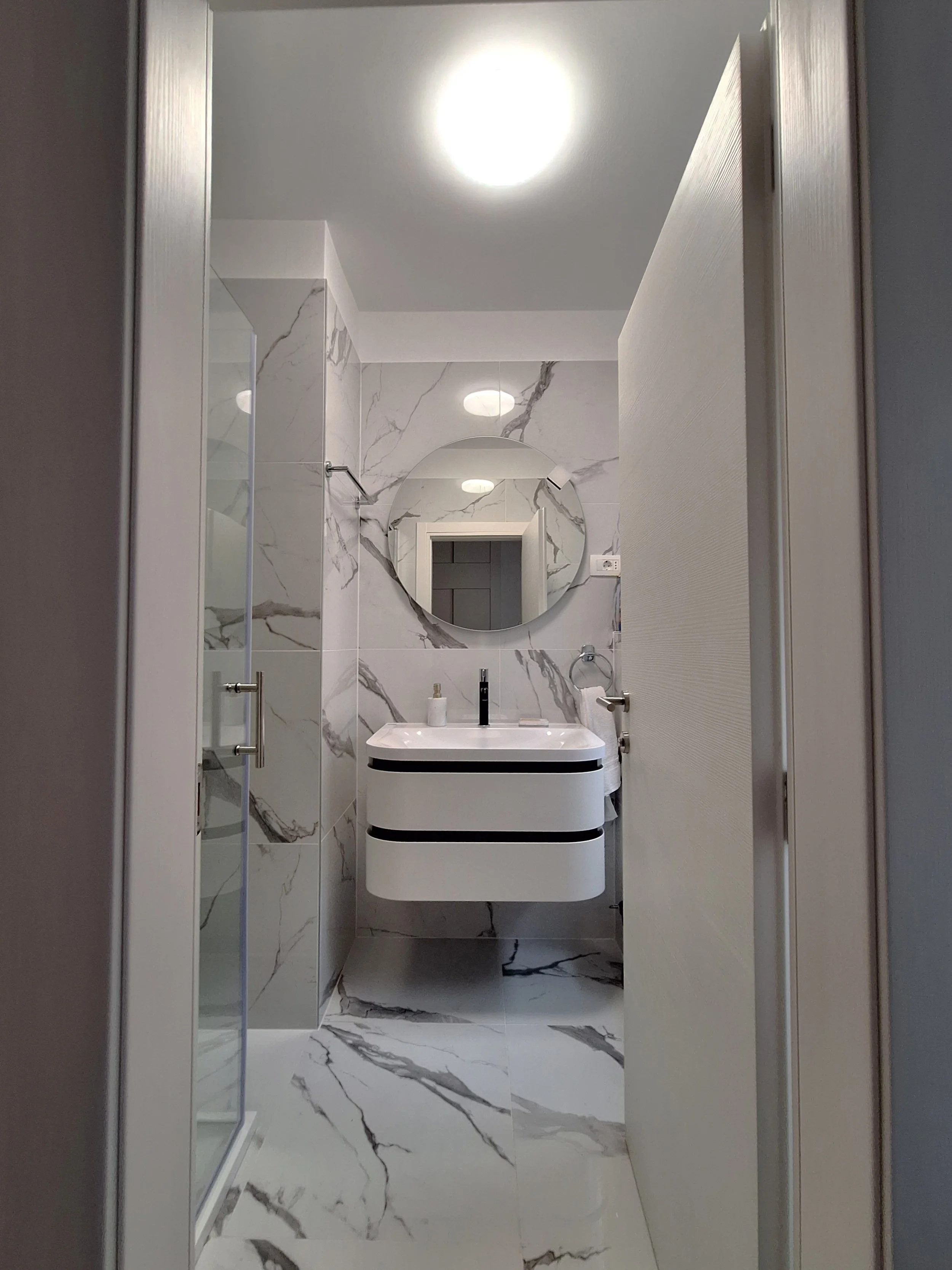 Modern bathroom with white marble walls and floor, a round mirror above a floating white vanity, and a glass shower door on the left.