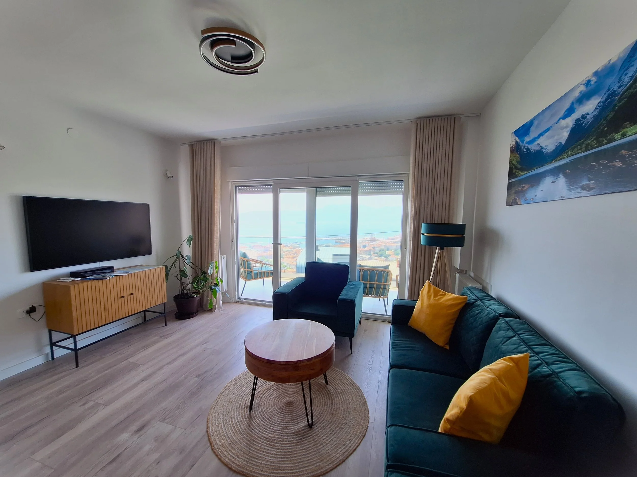 Living room with a dark green sofa, yellow pillows, a round wooden coffee table on a circular rug, a black armchair, a TV on a wooden stand, a large sliding glass door leading to a balcony with outdoor chairs, and a mountain landscape picture on the wall.
