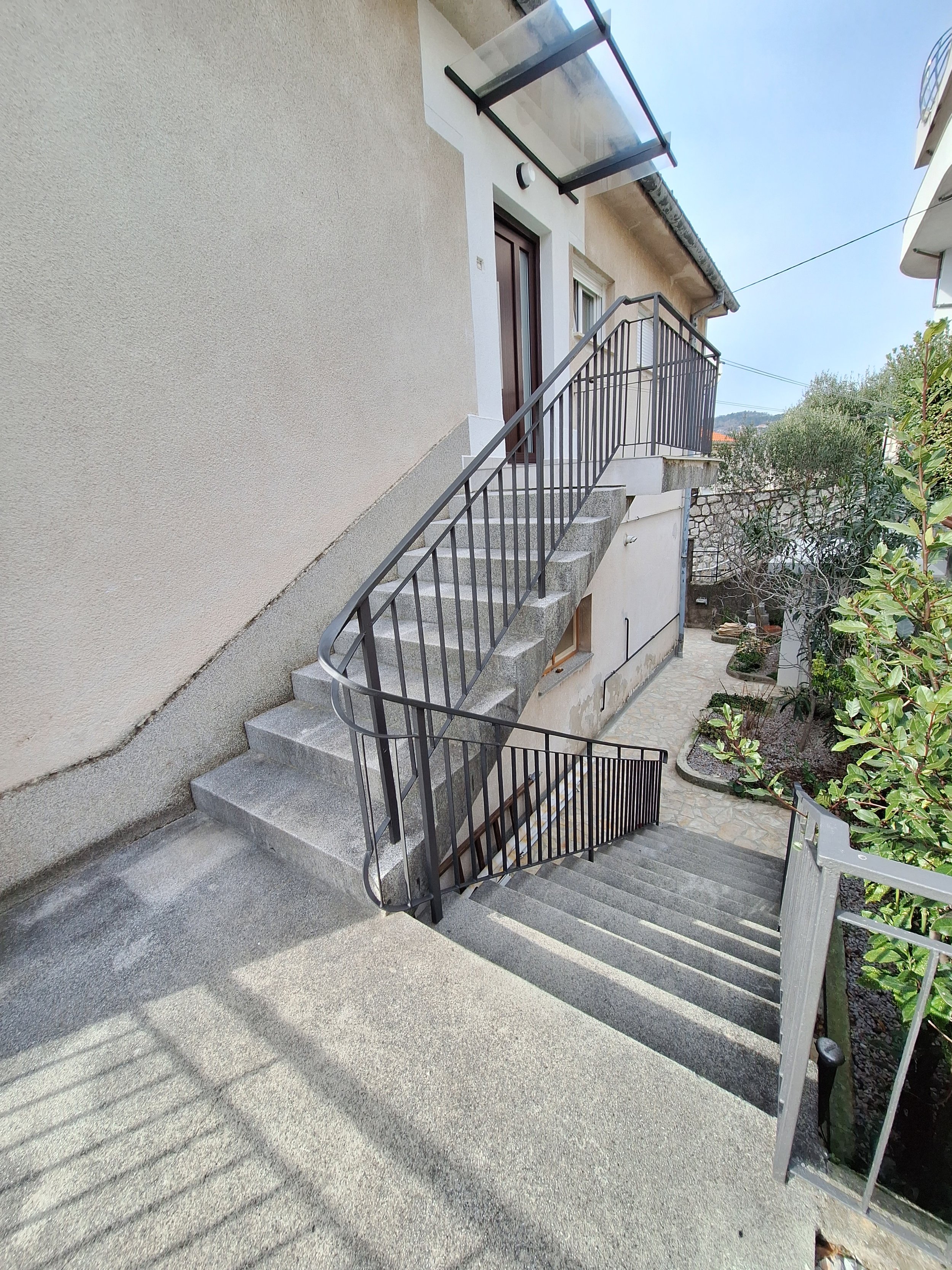 Exterior view of a house with a staircase leading to a door on the upper floor. The staircase has black metal railings and concrete steps. There is a small garden area with plants on the right side and a stone pathway.