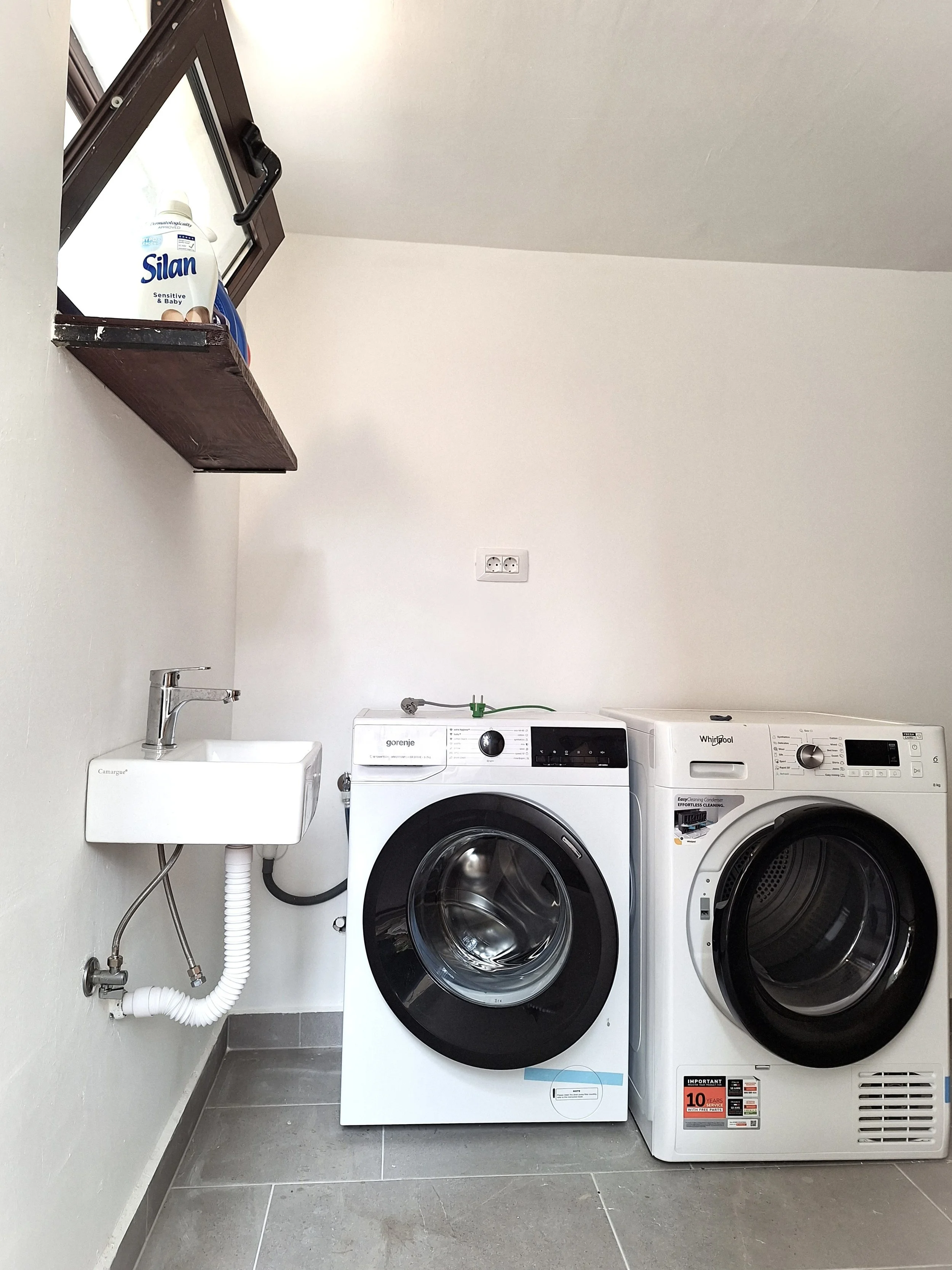Laundry room with a small sink, washing machine, and dryer. There is a window with a shelf holding a bottle of Silan fabric softener.