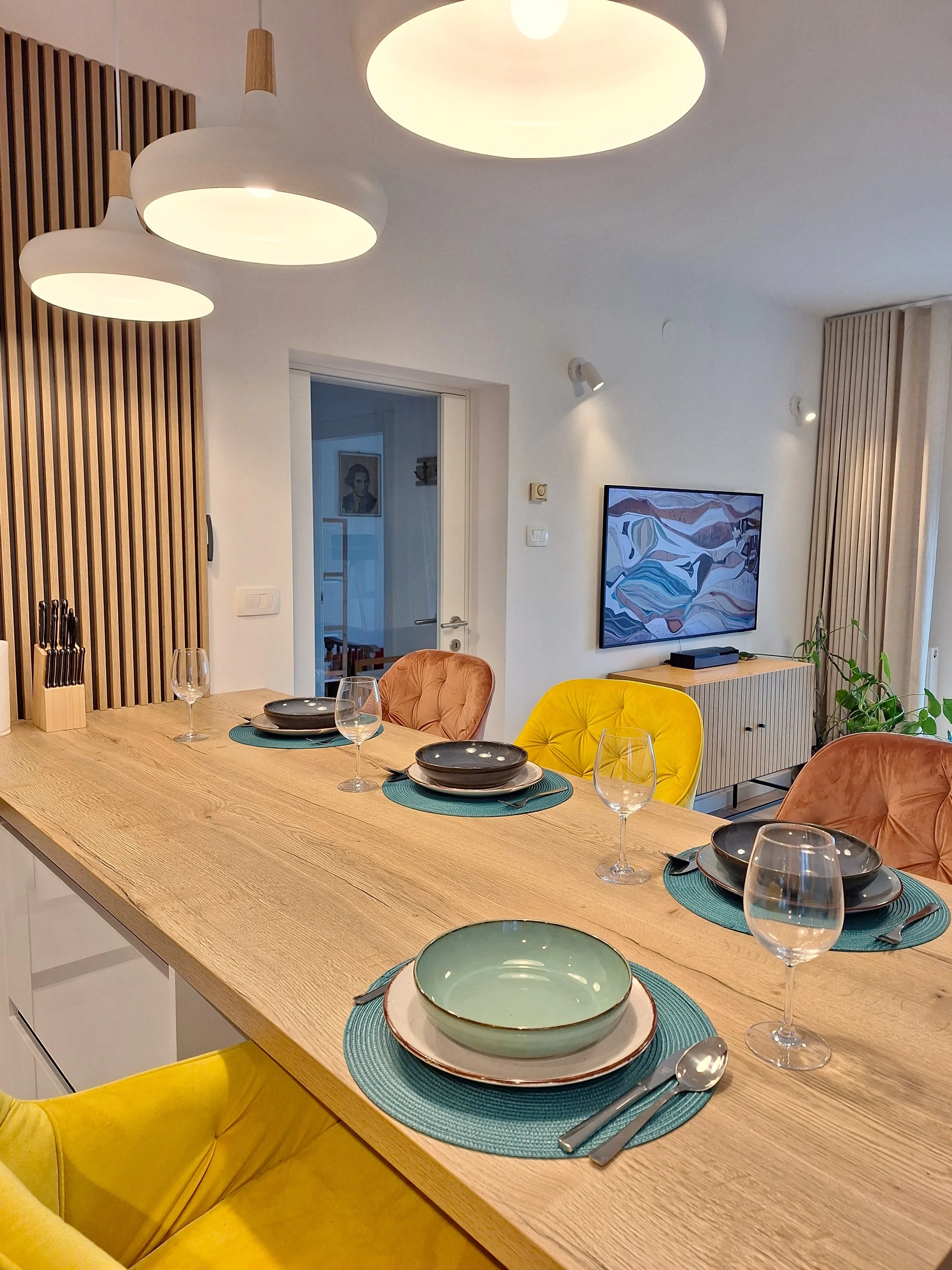 Dining room with a wooden table set with bowls, plates, wine glasses, and cutlery, surrounded by colorful upholstered chairs, modern pendant lights, and a wall-mounted TV.