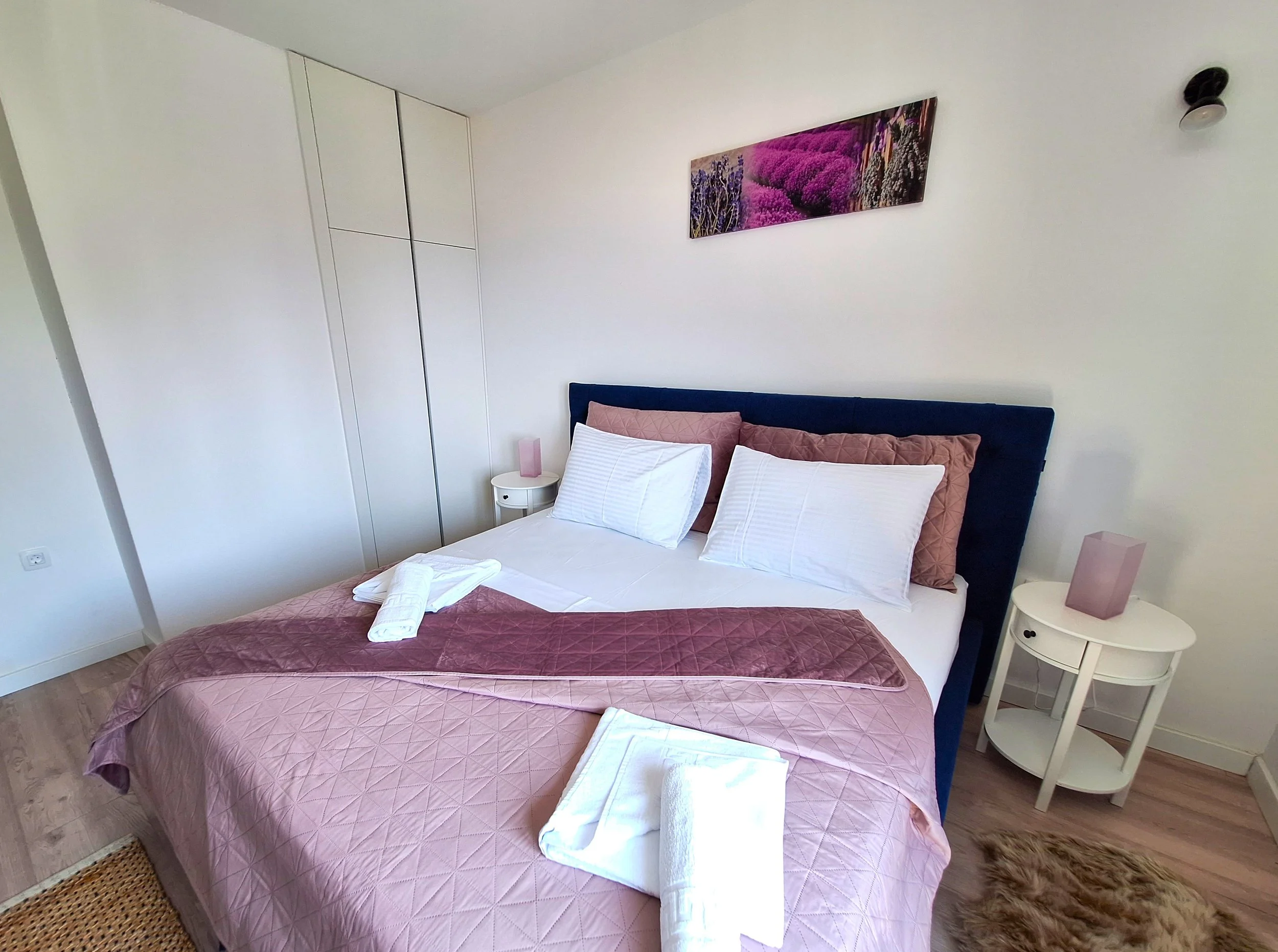 A neatly made bed with pink and white bedding, two white pillows, and a dark blue headboard in a minimalist bedroom. There are two nightstands with pink lamps and a picture of lavender flowers on the white wall.