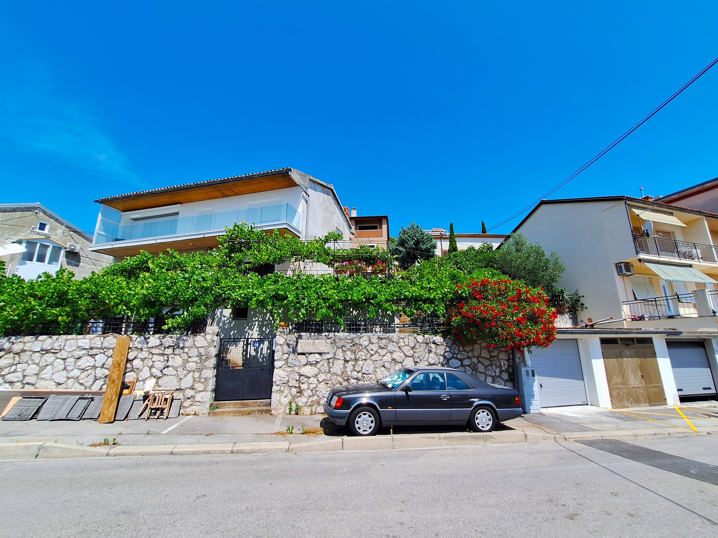 Residential hillside with modern houses, greenery, and a black car parked on the street under a bright blue sky.