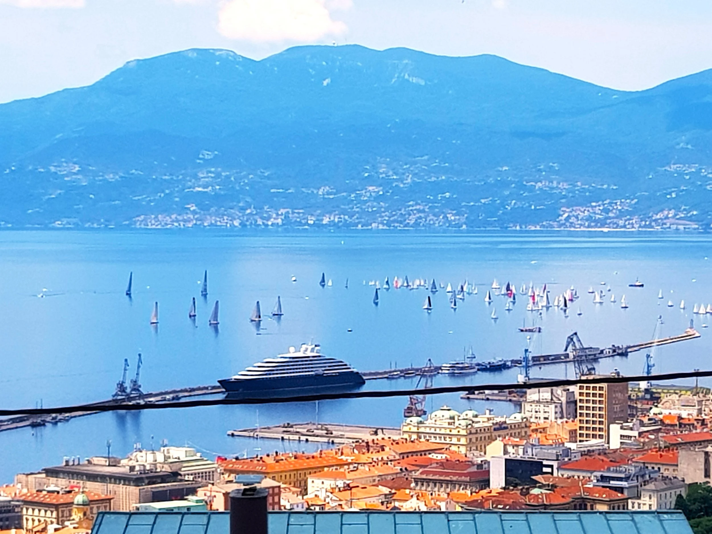 View of a harbor with numerous sailboats and yachts, a city with red-tiled roofs, and mountains in the background.