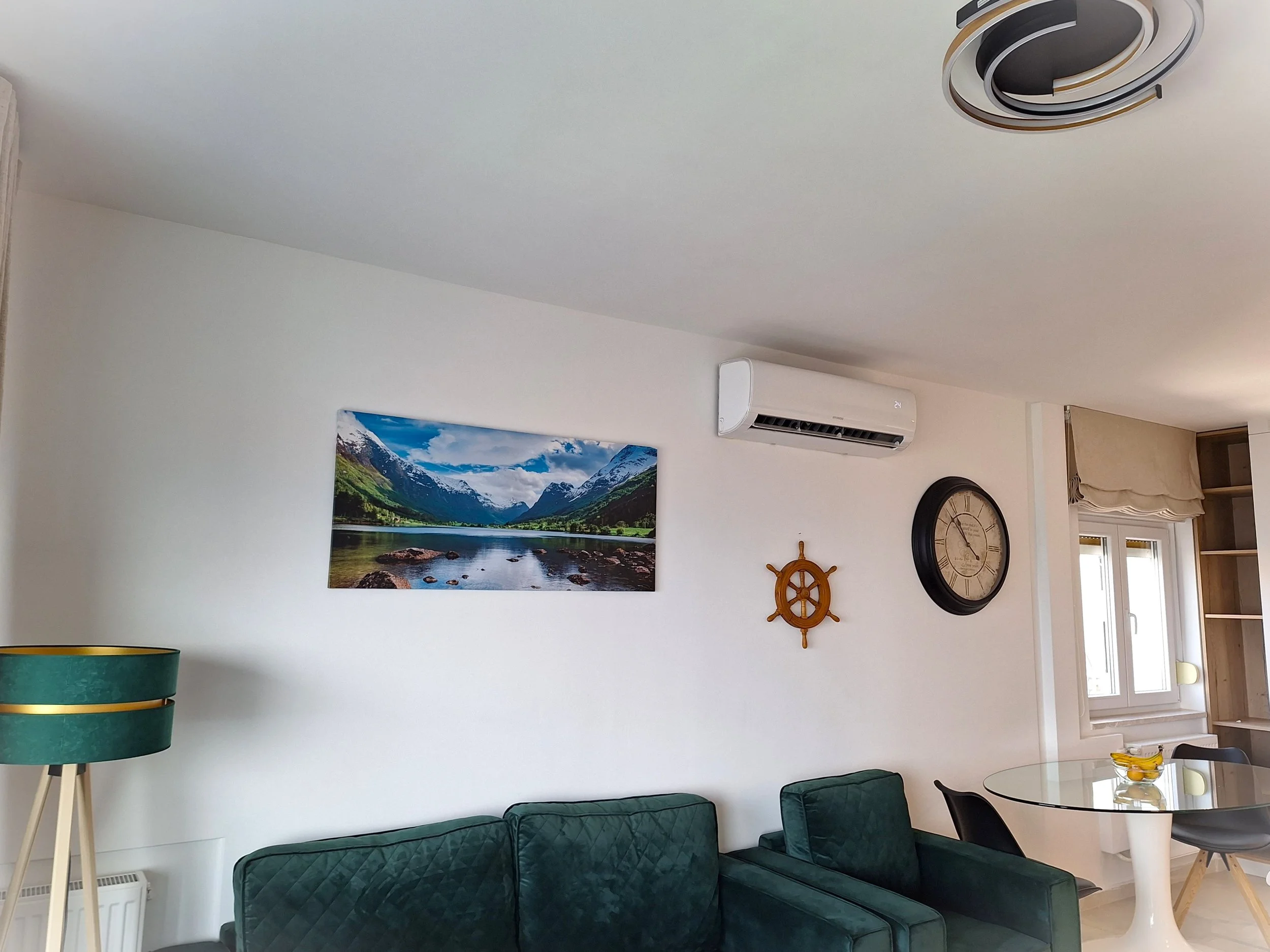 Living room with a white wall, green sofa, a landscape photo of mountains and a lake, a wall clock, a nautical ship wheel, air conditioning unit, a round glass dining table with chairs, a window with beige curtains, and a floor lamp with a green shade.