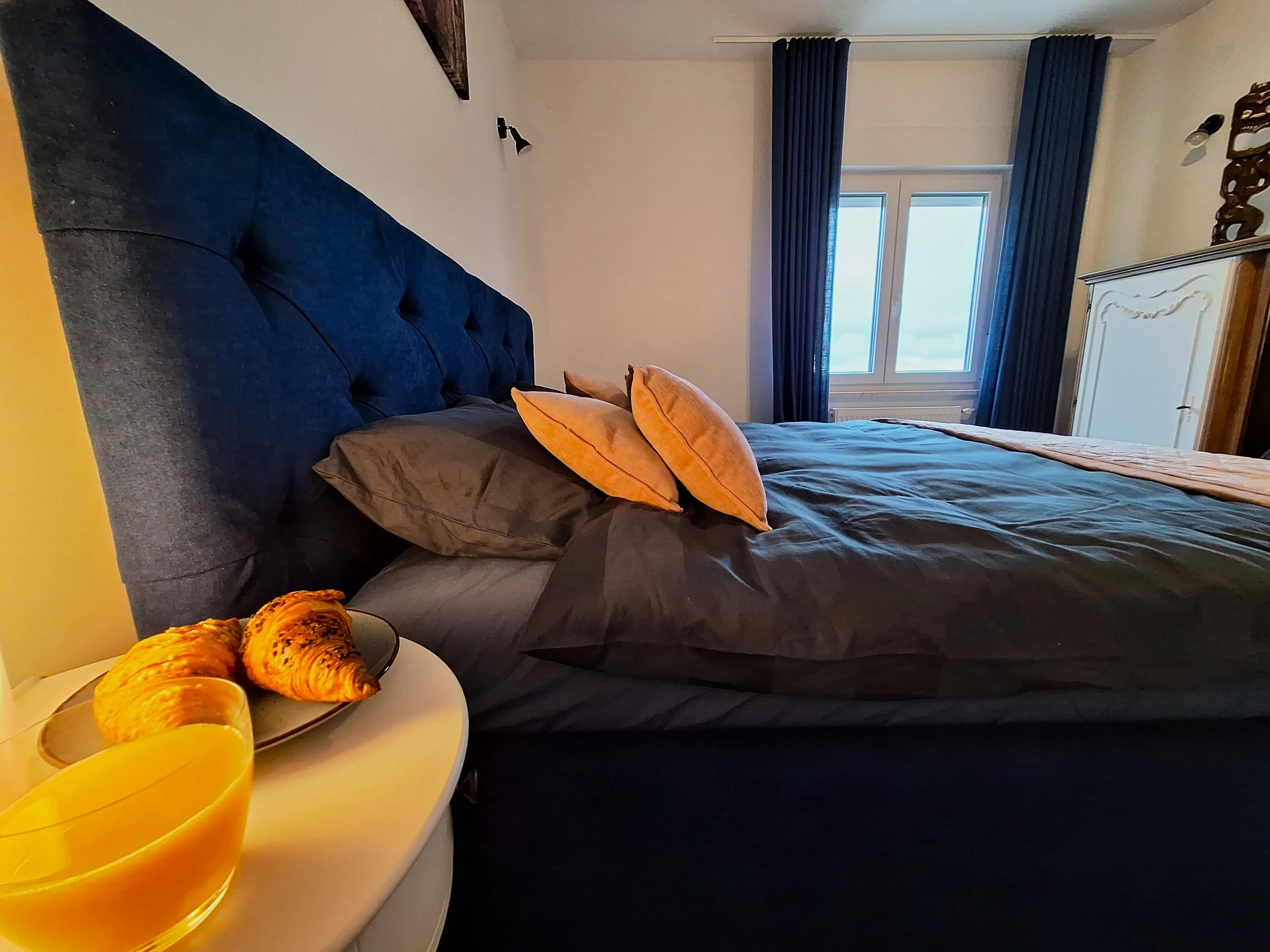 A bedroom with a large gray bed, orange accent pillows, and dark blue curtains over a window. A white nightstand with croissants and orange juice is beside the bed. An ornate white wardrobe is in the background.