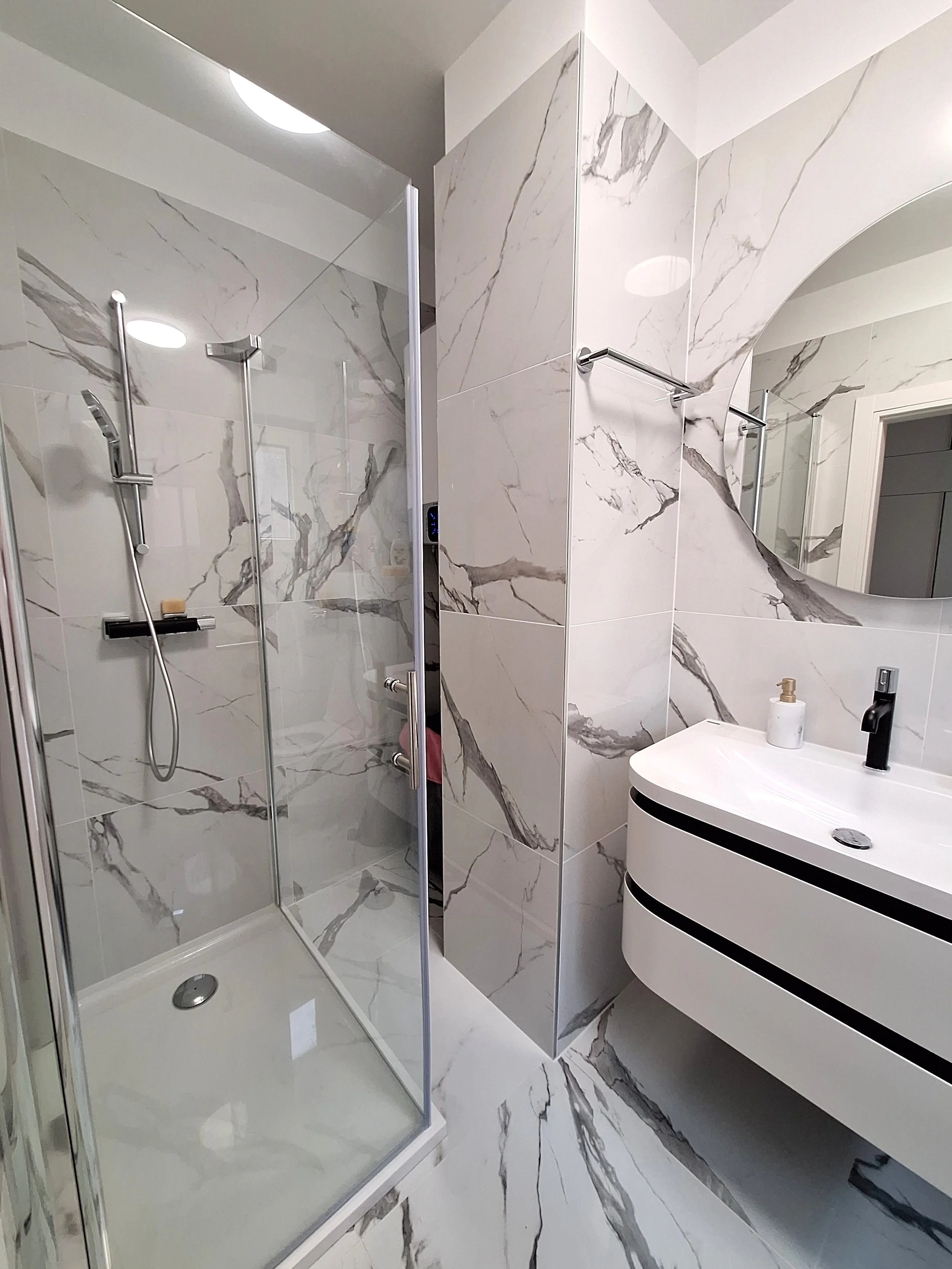 Modern bathroom with marble walls and flooring, a glass shower enclosure, and a white vanity with a black faucet and soap dispenser.