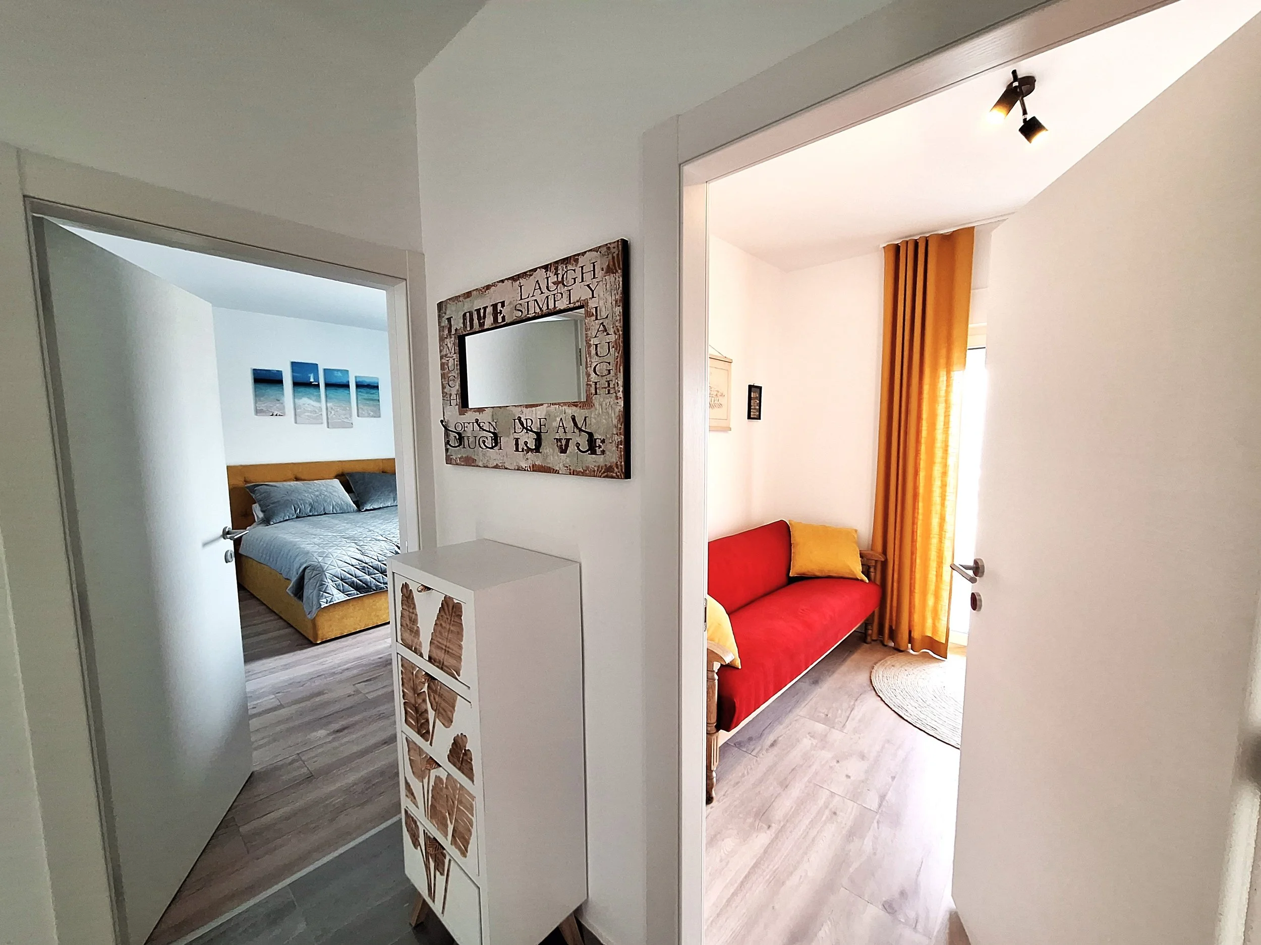 Interior view of a hallway leading to two bedrooms, one with a red couch and yellow curtains, and the other with a bed and three wall art pieces.