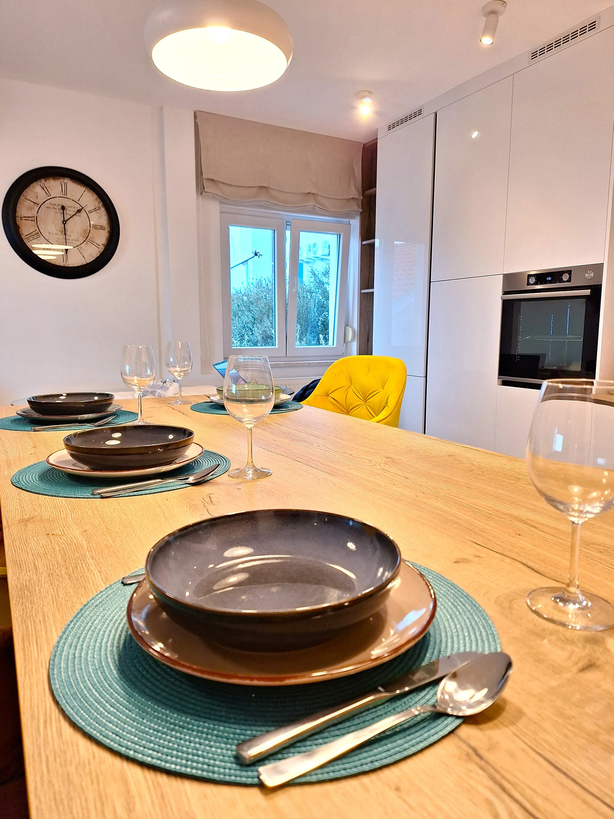 A dining table set for a meal with black bowls, white plates, wine glasses, and silverware on turquoise placemats, in a modern kitchen with white cabinets, a yellow chair, and a clock on the wall.