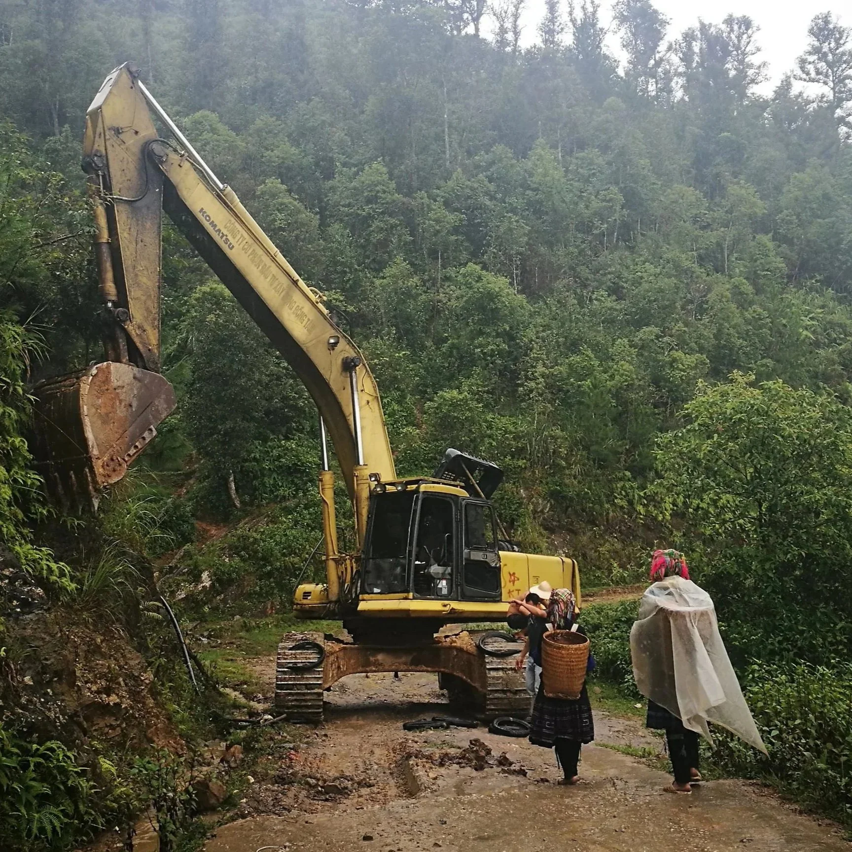 An excavator working on a mountain road, with two women walking nearby, one carrying a basket and the other covered with a plastic sheet, amidst lush green forest.
