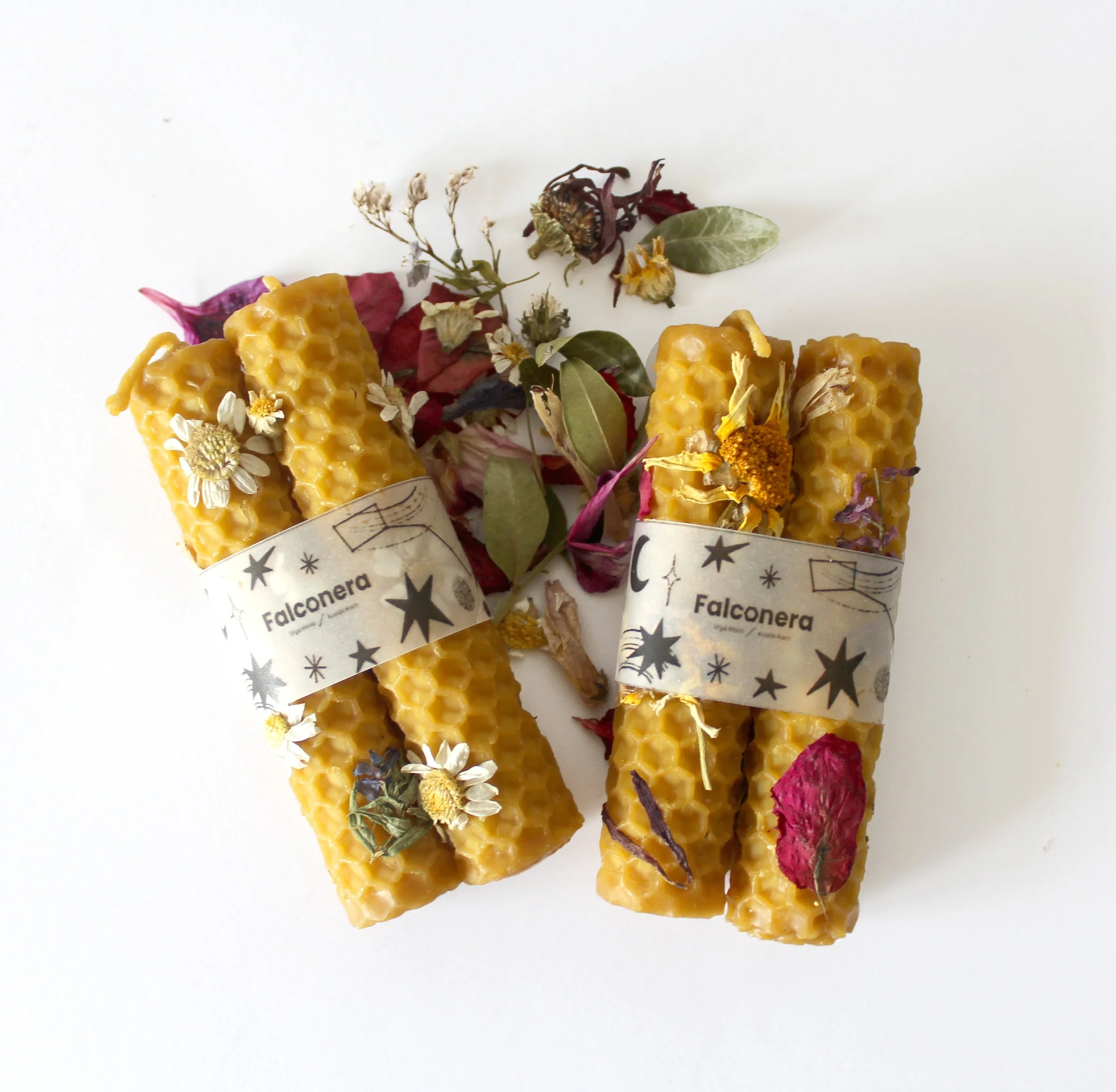 Dos paquetes de velas de cera en forma de panel con etiqueta que dice 'Falconera', rodeadas de flores secas y hojas en un fondo blanco.