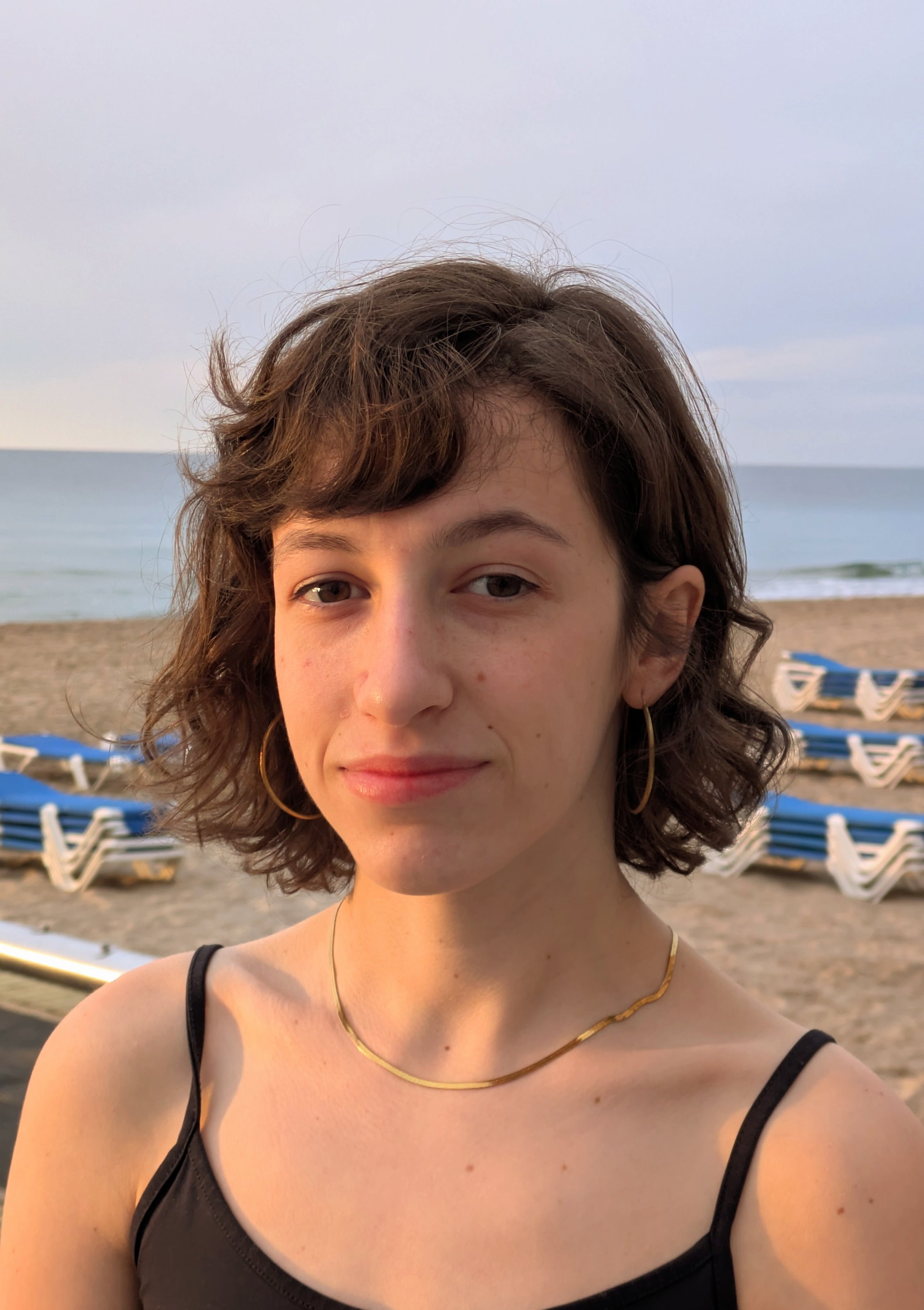 Retrato de una joven con cabello rizado en la playa, con sillas y el mar en el fondo.