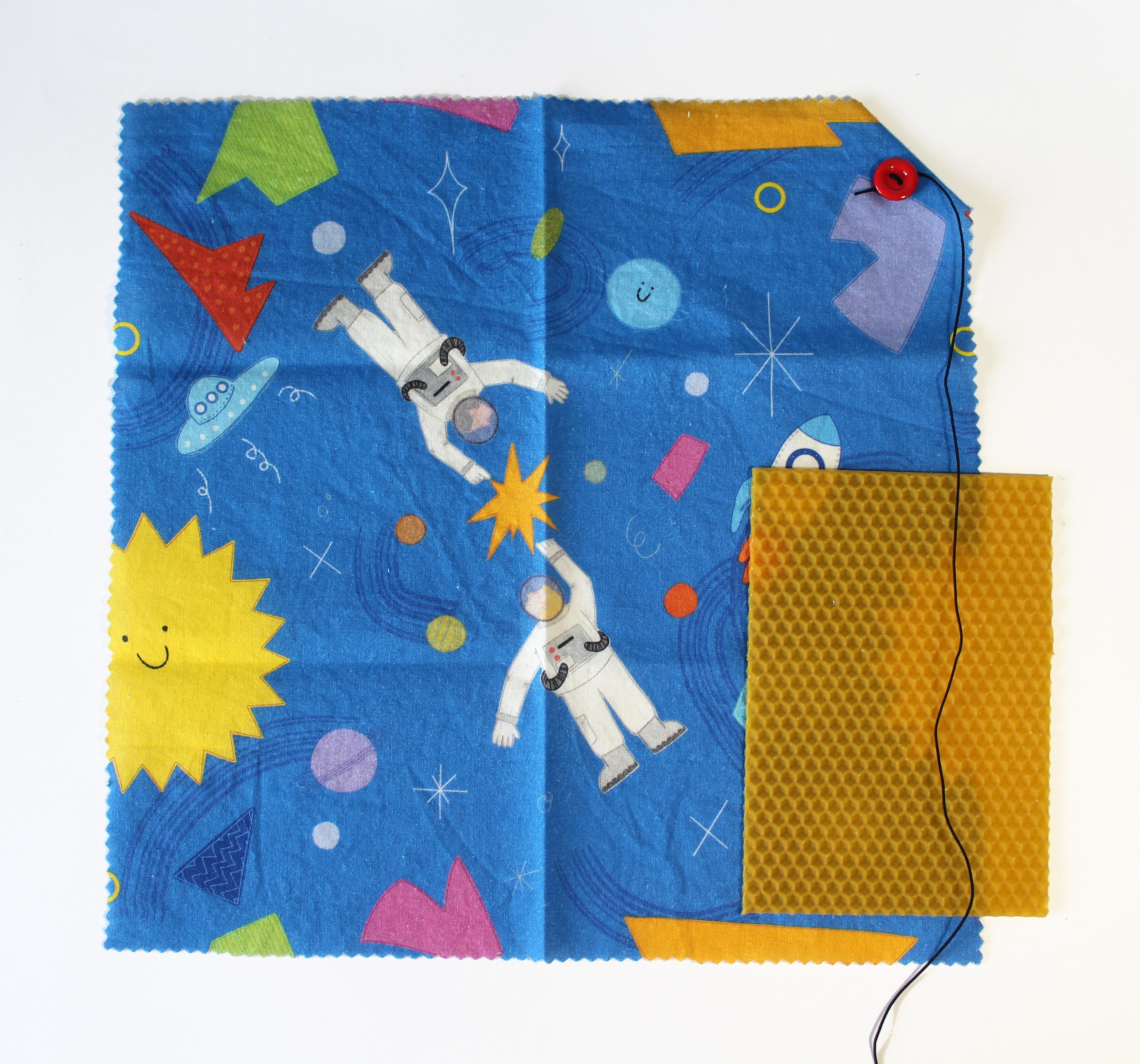 Tela de tela de mascota con un fondo de espacio. Tiene dibujos de astronautas, un sol sonriente, planetas y estrellas. Incluye una pequeña bolsa de malla amarillo y un botón rojo con hilo negro.