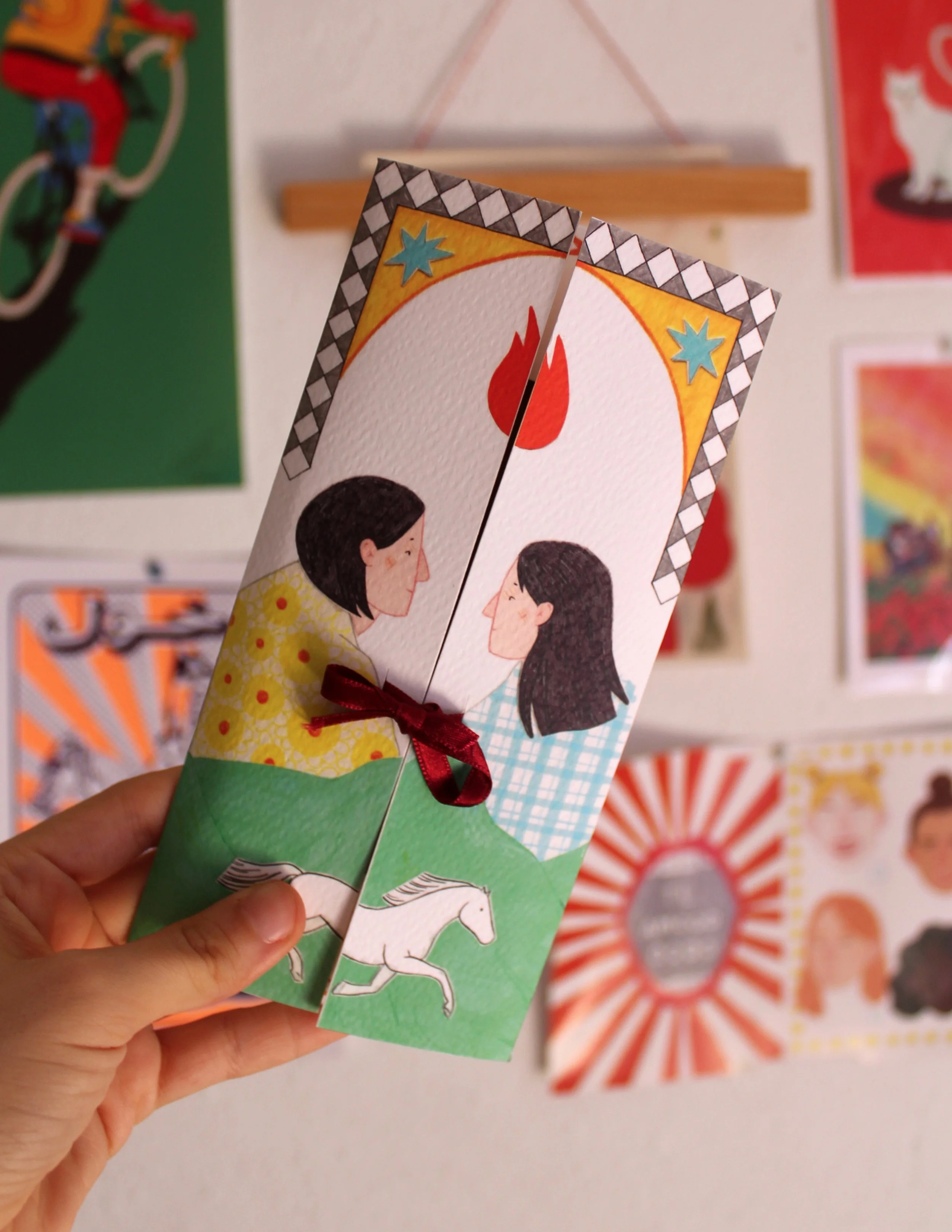 Tarjeta decorativa con ilustración de dos mujeres mirándose, una con cabello negro y otra con cabello castaño, con un símbolo de fuego entre ellas y caballos en la parte inferior, sujetada con una cinta de tela. En el fondo, varias carteles y arte en