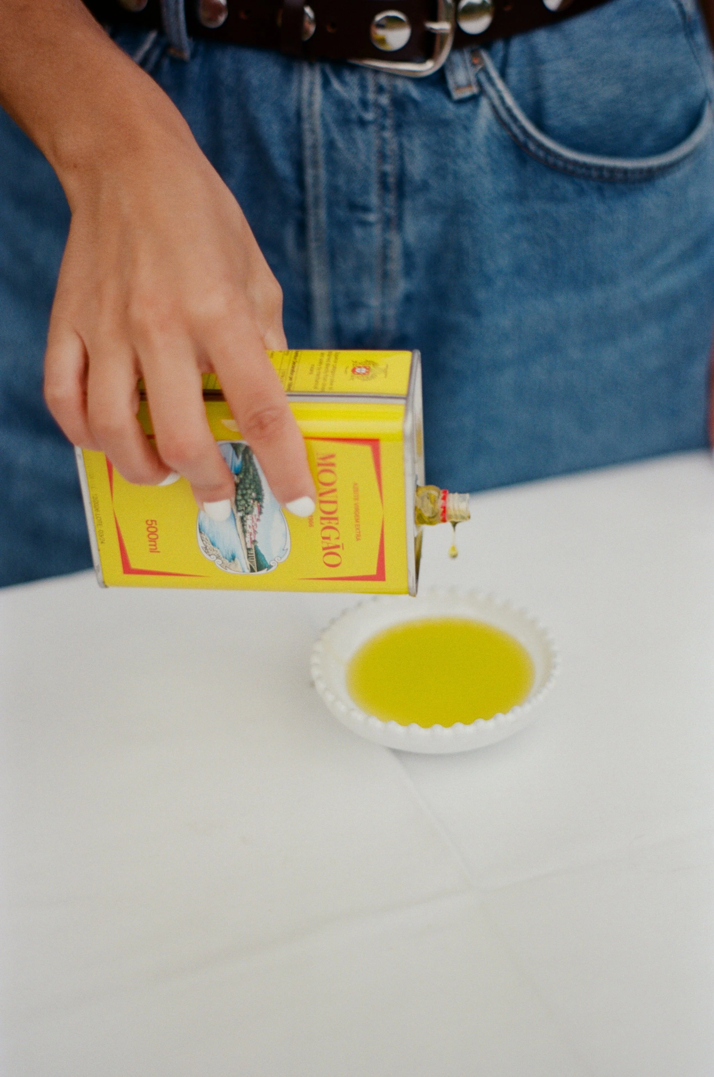 Person pouring a yellow bottle of olive oil into a white bowl on a white table.
