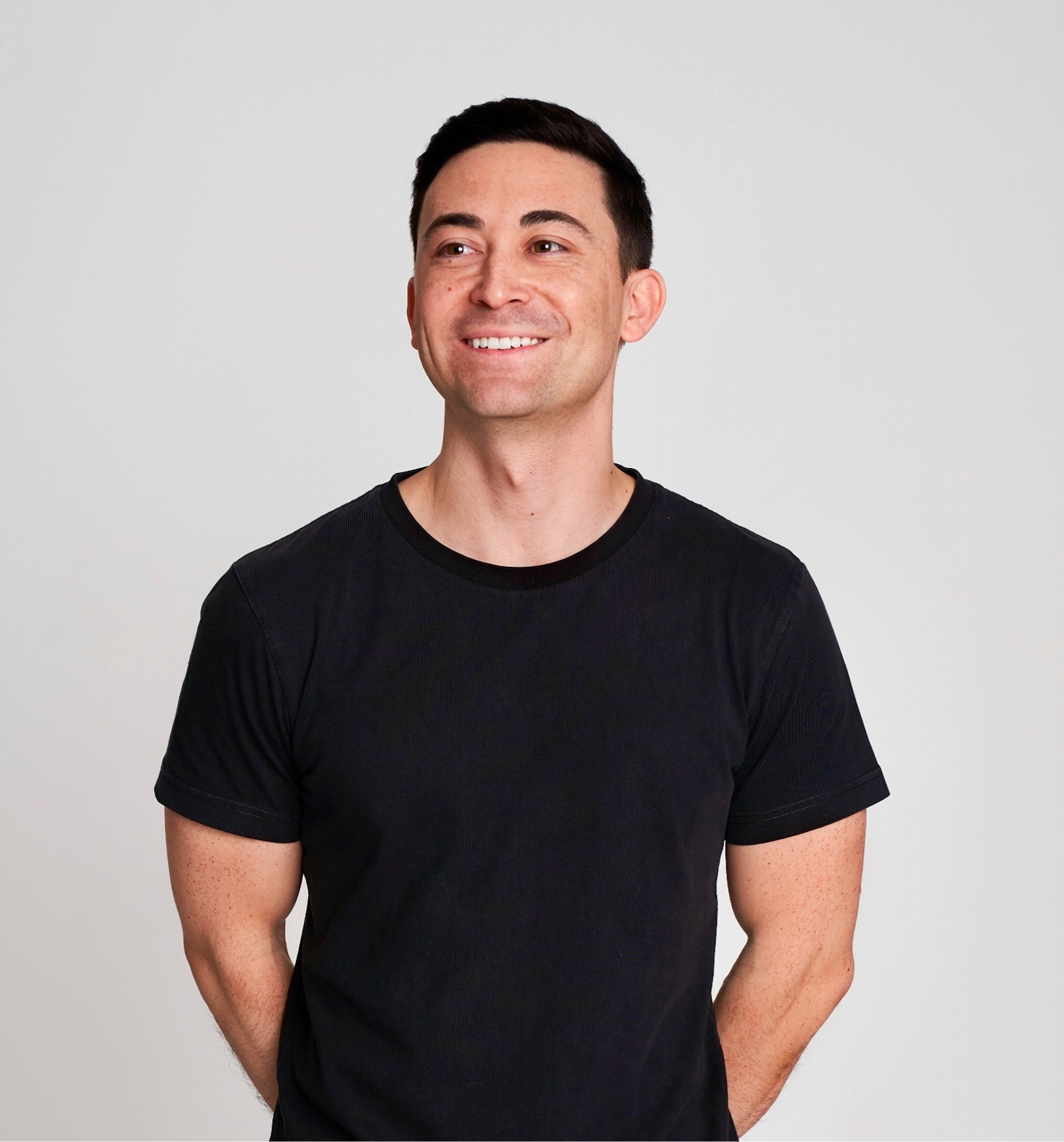 A smiling man with short dark hair wearing a black T-shirt standing against a plain light gray background.