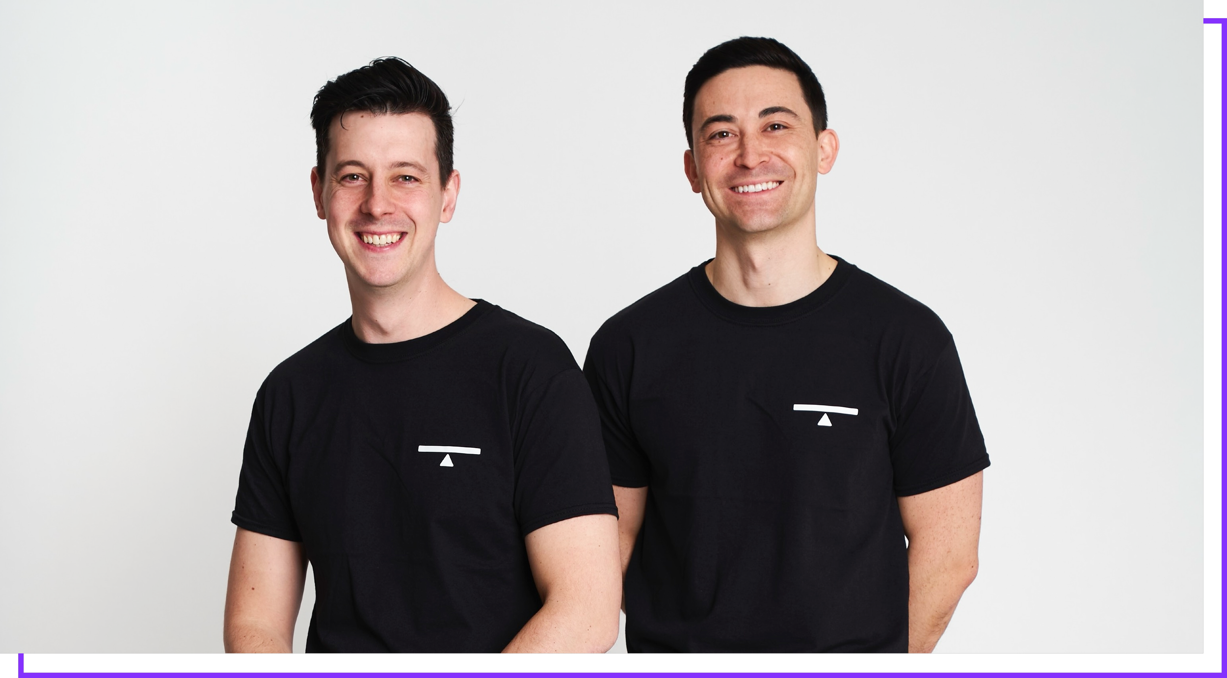 Two men standing side by side, smiling, wearing black t-shirts with a small white logo on the left chest, against a plain white background.
