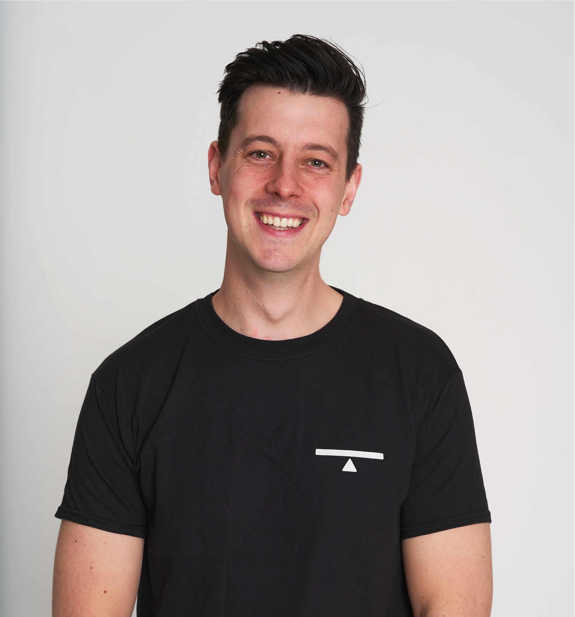 A young man with dark hair smiling, wearing a black T-shirt with a small white geometric logo on the chest, against a plain white background.