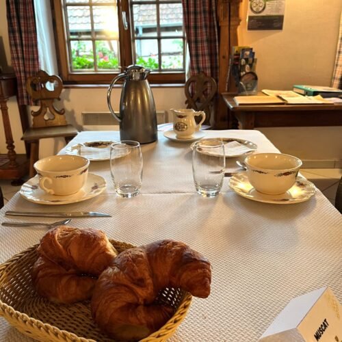 Een ontbijttafel met drie kopjes, drie glazen, een kan, een melkpafkaardje en twee croissants in een mand bij het chateau van Ruhlmann-Schutz