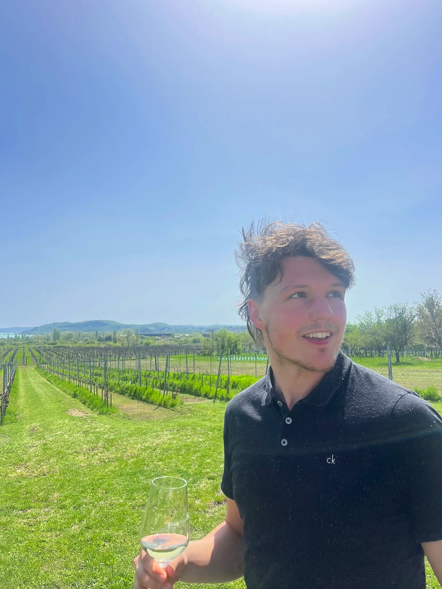 Eigenaar van Vino by Palazzo met een glas wijn in de hand op een Hongaarse wijngaard op een zonnige dag, blauwe lucht, groene velden en wijnranken op de achtergrond.