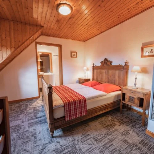 Een gezellige slaapkamer met een houten bed, rood en wit beddengoed, twee nachtkastjes met lampen, een muur met foto's, en een plafond van hout bij het chateau van Ruhlmann-Schutz