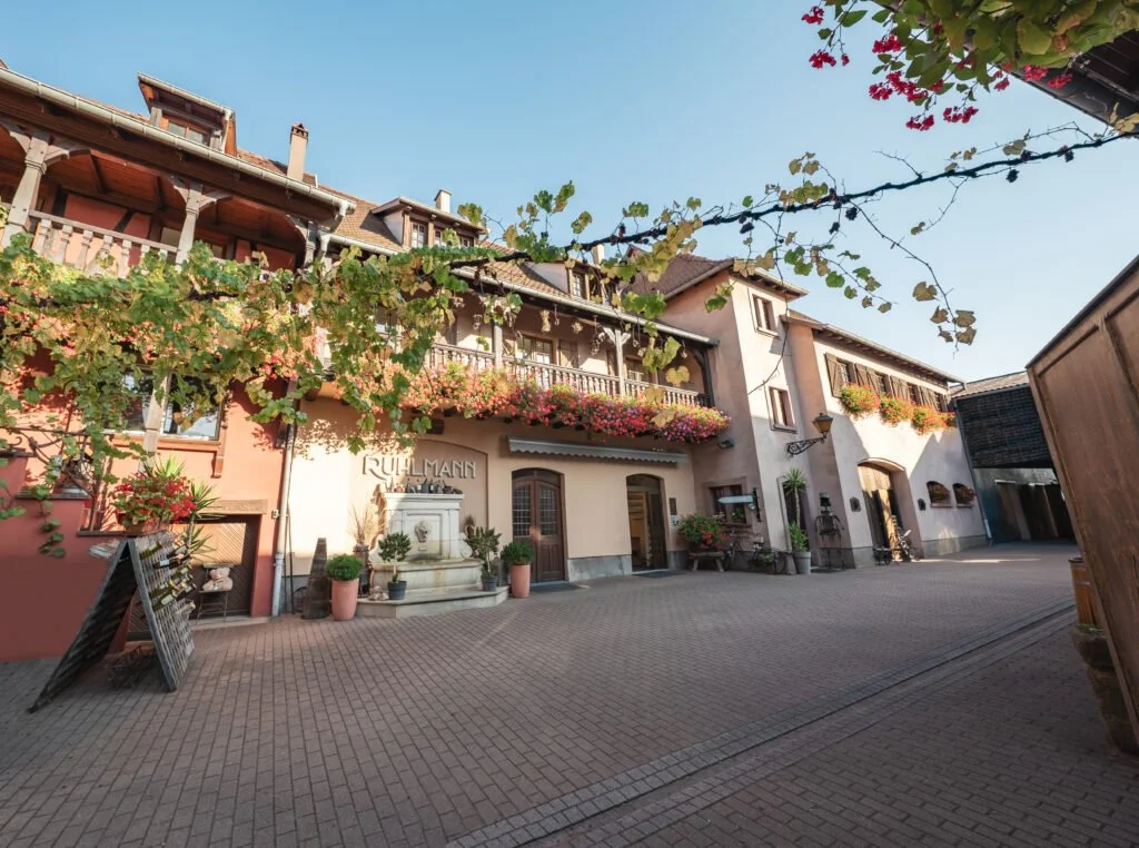 Een charmant traditioneel gebouw met bloemen en planten, een bron, op een rustige plein onder een blauwe lucht. bij het chateau van Ruhlmann-Schutz