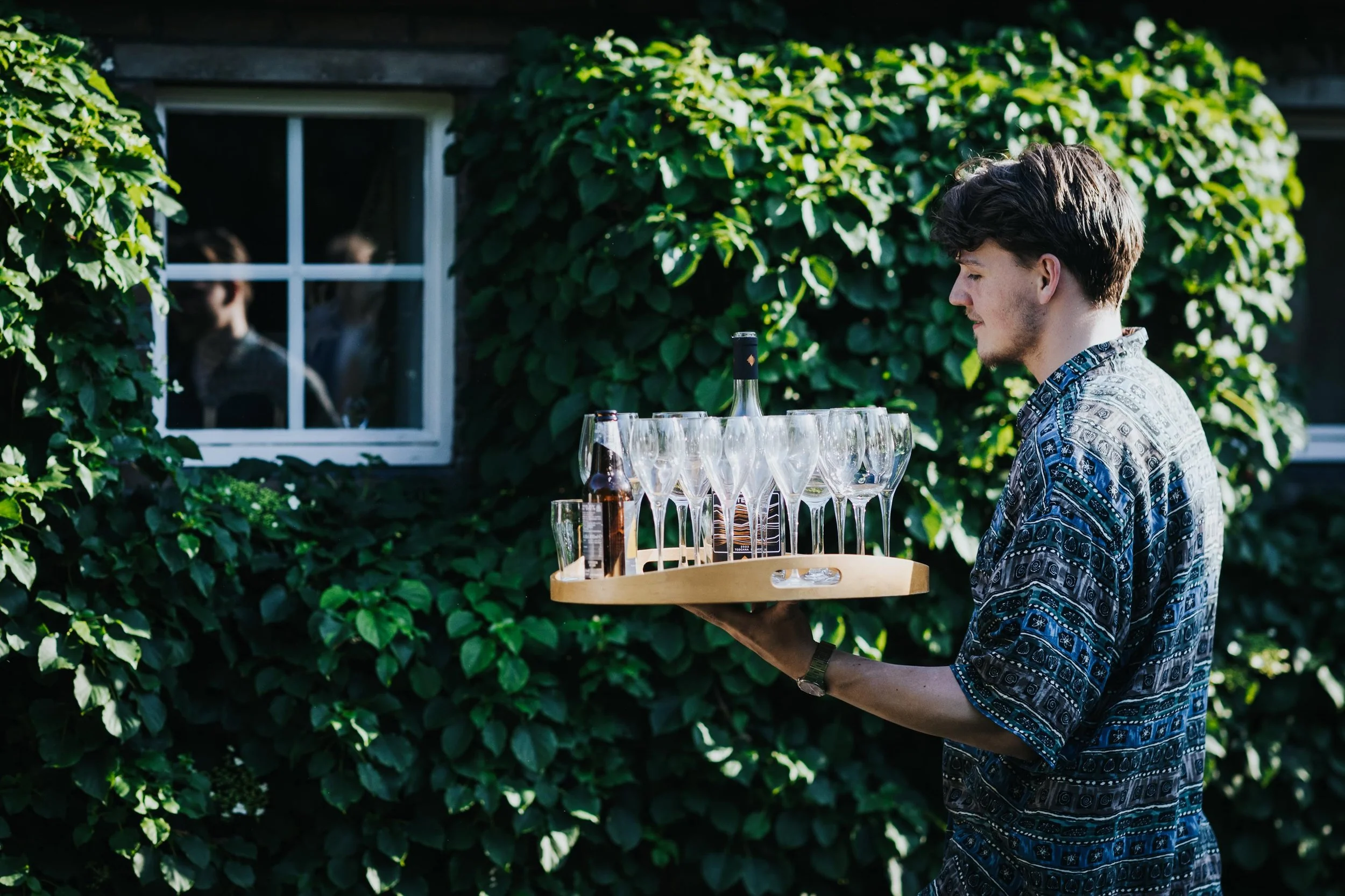 Jonge man draagt een houten dienblad met lege champagneglazen en een wijnfles, buiten bij een groene muur met raam.