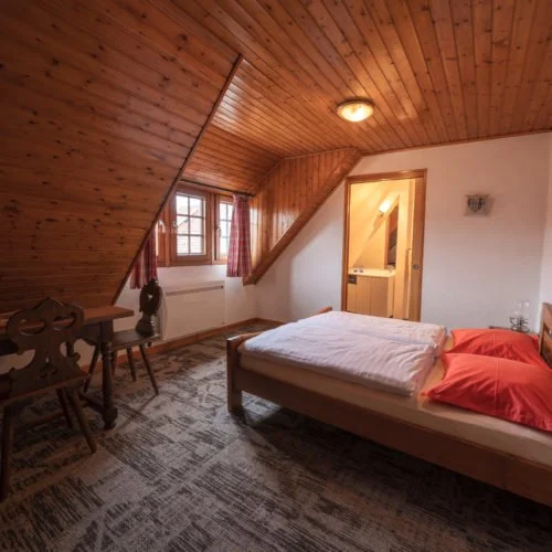Slaapkamer met een bed, raam met rood geruite gordijnen, een houten plafond en muur, een mandje en een tafel met stoelen, en een open deur naar een kleine badkamer bij het chateau van Ruhlmann-Schutz.
