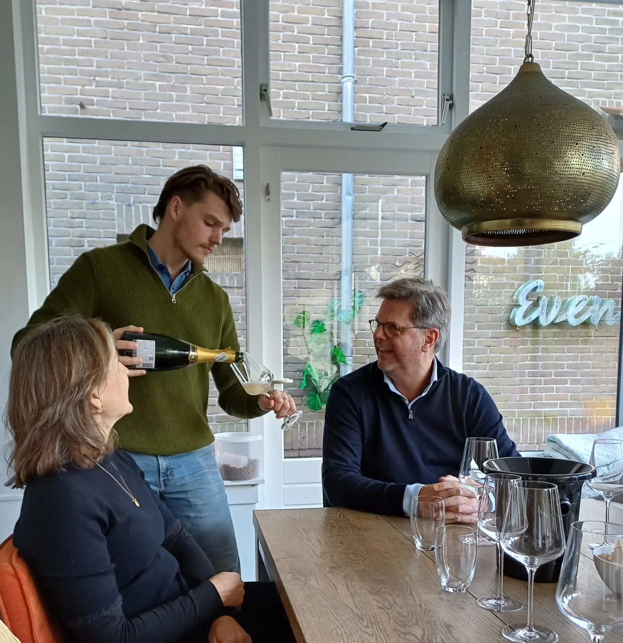 Drie mensen genieten van wijn in een goed verlichte kamer met grote ramen en een houten tafel. Een man schenkt wijn in een glas terwijl een vrouw en een man met een lach naar hem kijken.