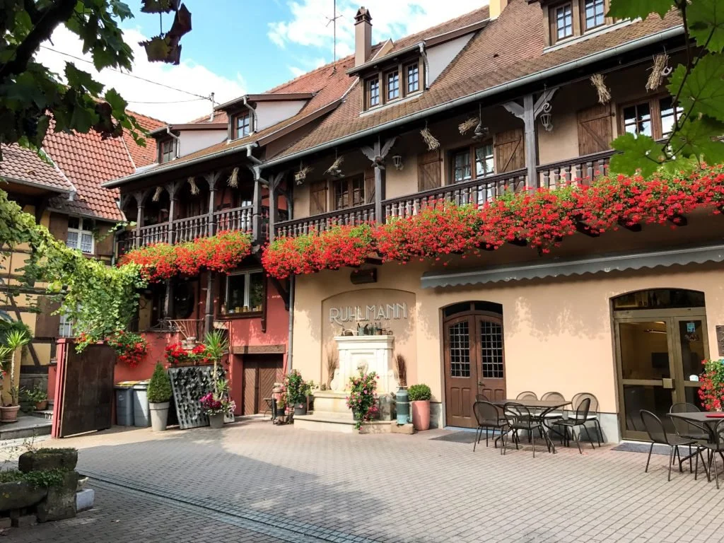 Een historische gevel met een overdekte balkon vol roze bloemen, terras met tafels en stoelen, en een ingang met een bord 'Ruhmann' bij het chateau van Ruhlmann-Schutz