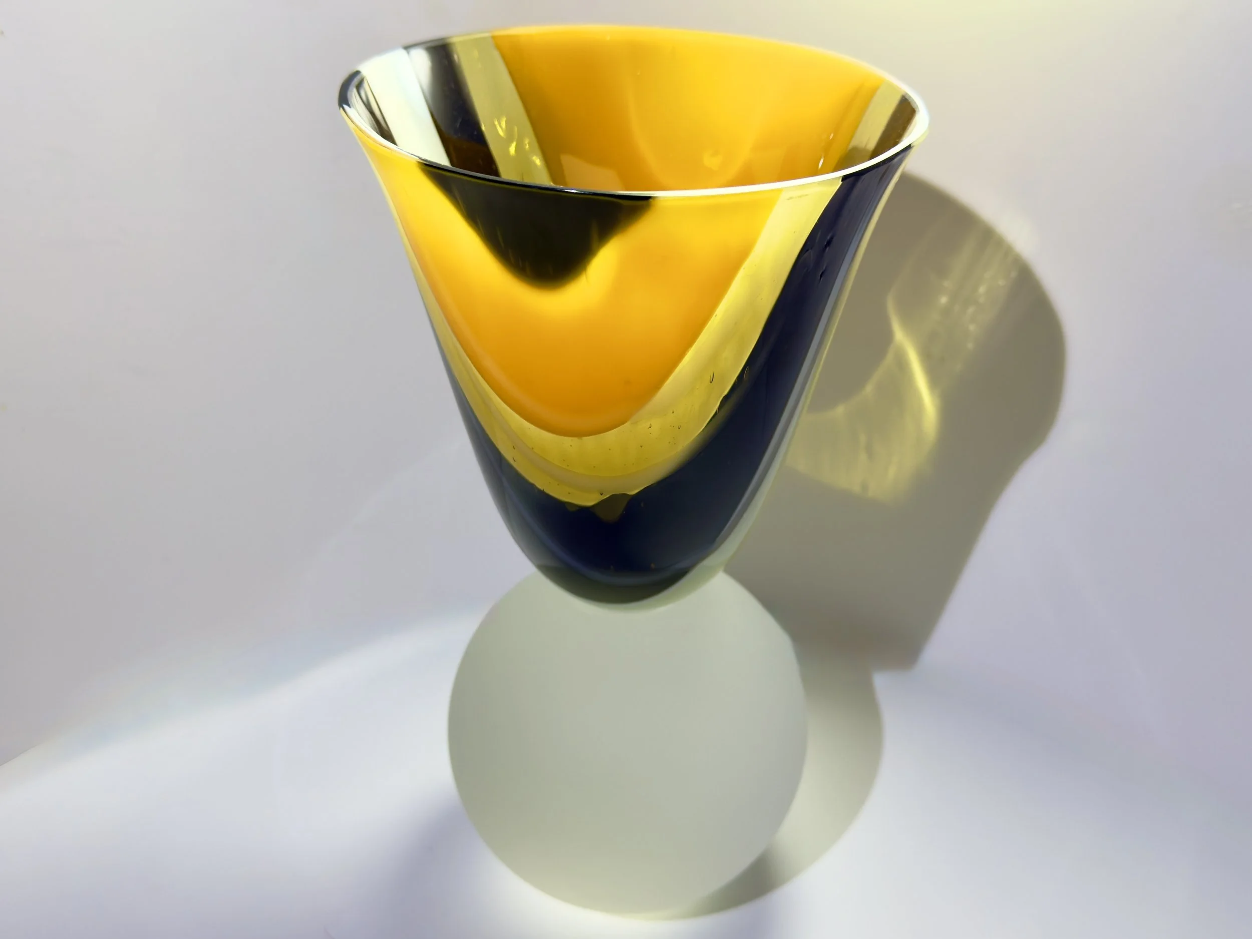A colorful glass vase with yellow, black, and clear sections, casting a shadow on a white surface.