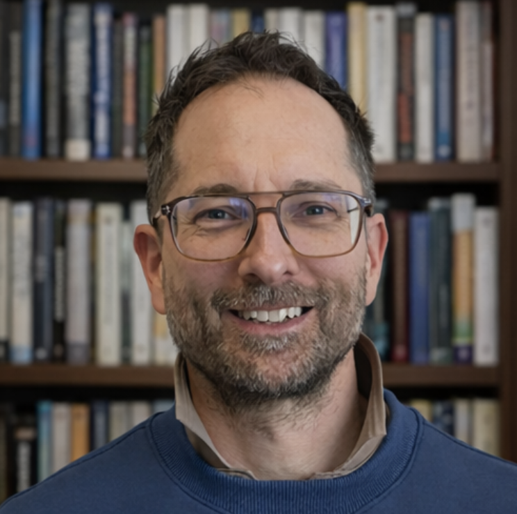 A smiling man with glasses and a beard standing in front of a bookshelf filled with books.