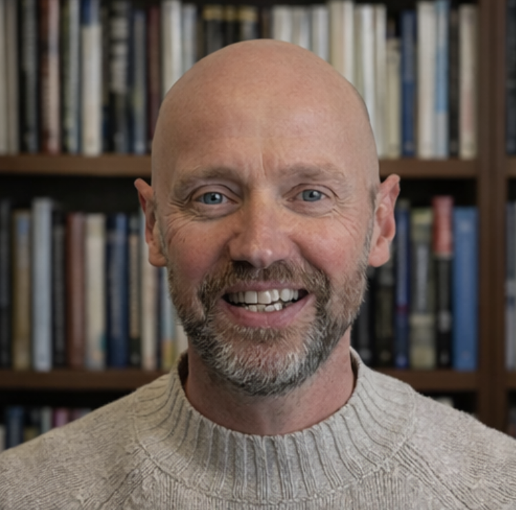 A smiling man with a beard and glasses, wearing a beige knit sweater, standing in front of a bookshelf filled with books.