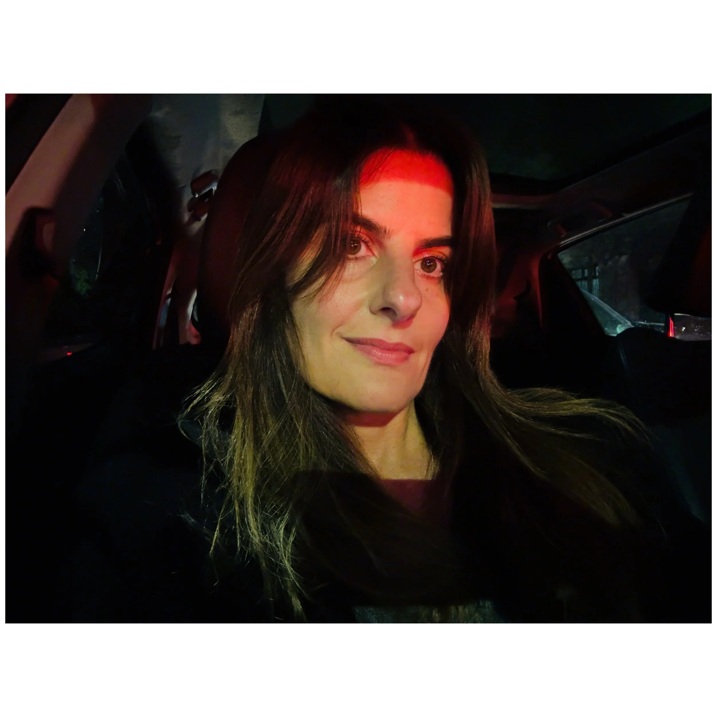 Close-up of a smiling woman with brown and blonde hair sitting inside a car at night, illuminated by red and green interior lights.