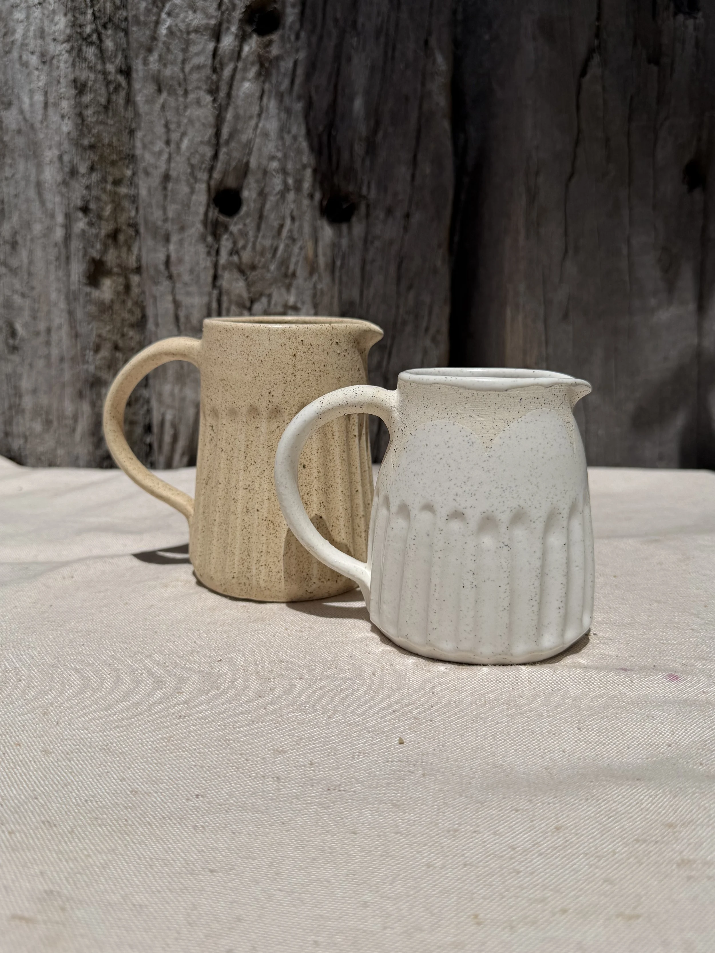 Two ceramic pitchers, one beige and speckled, the other white with textured vertical ridges, sit on a neutral-colored fabric surface with a weathered wooden background.