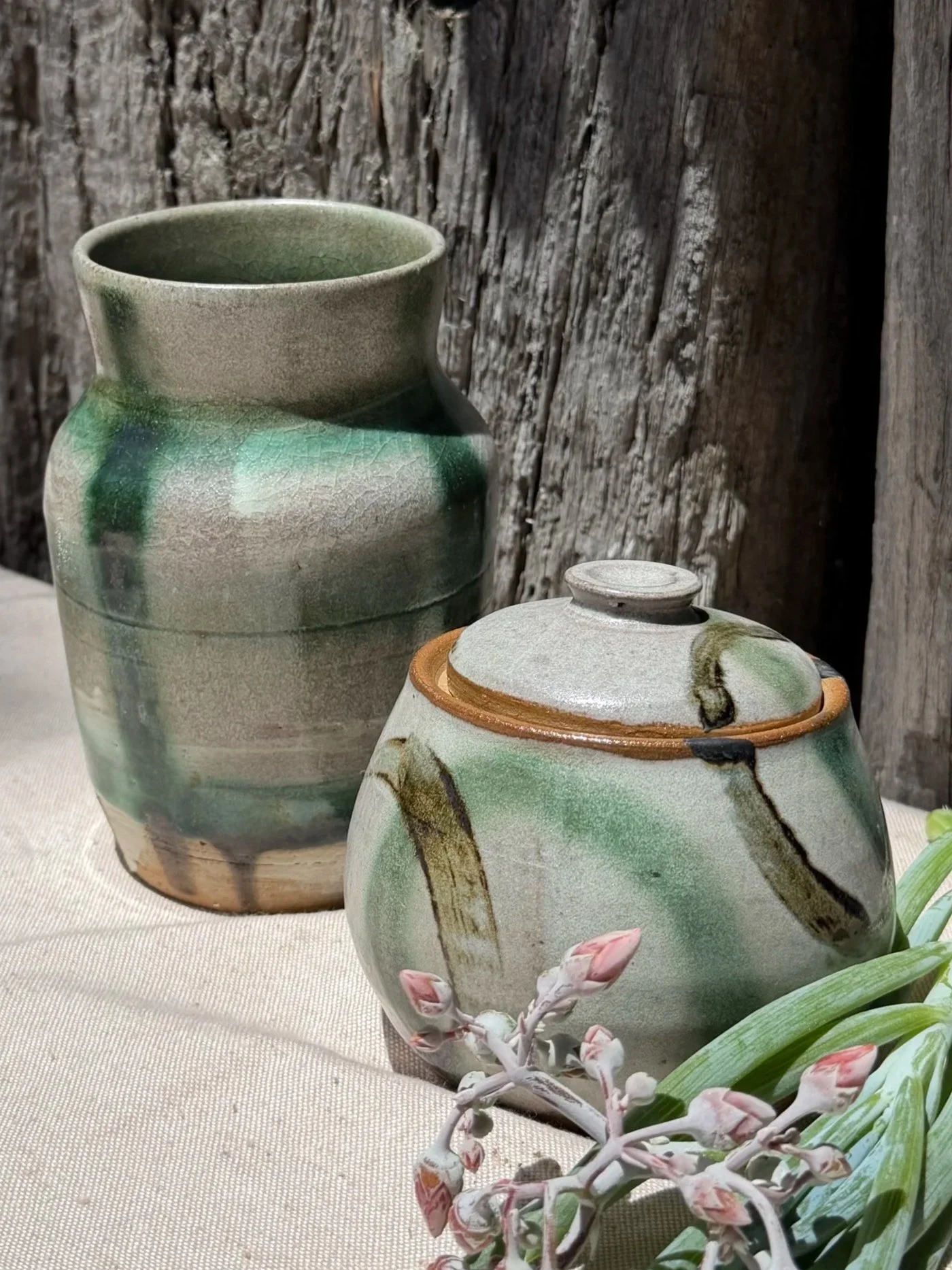Two ceramic vases with green, earthy glaze, placed on a cloth surface against a weathered wooden background. One vase has a lid, and there are pink flowers and green leaves in front of the vases.