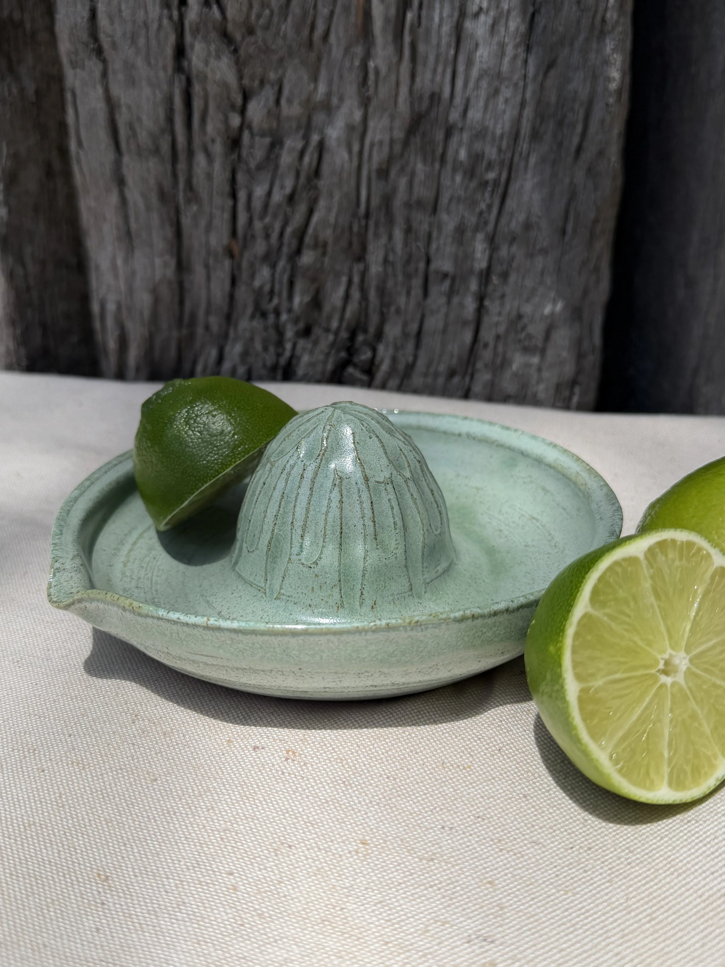 A green ceramic citrus juicer with half a lime on top and a whole lime in front, placed on a light-colored cloth with a wooden background.