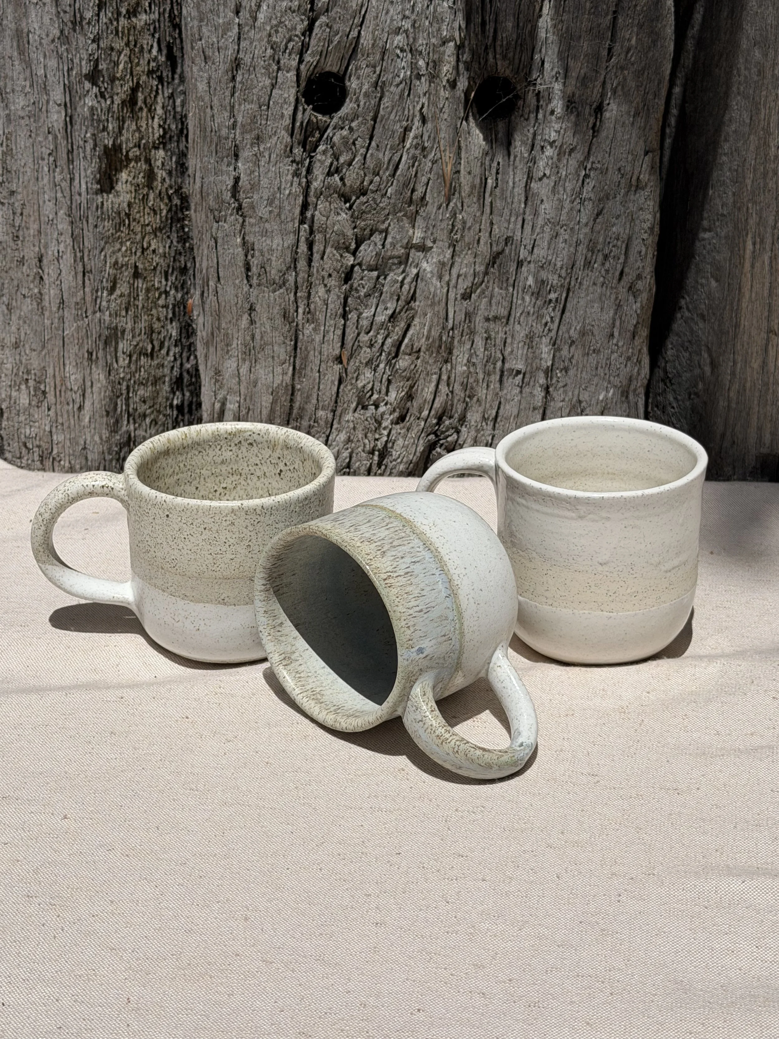 Three beige ceramic coffee mugs with speckled glaze on a light fabric surface, with a weathered wooden background.