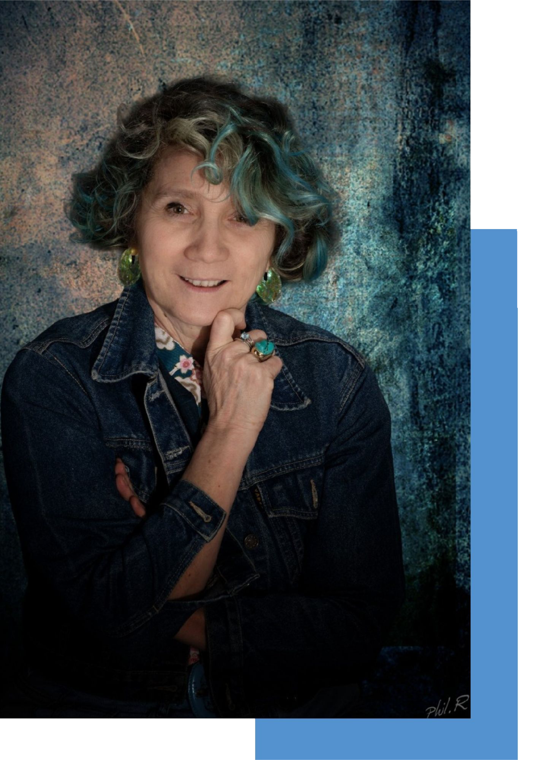 Portrait de Véronique Gillet-Chevais femme avec des cheveux bouclés bleu, portant une veste en jean et des boucles d'oreilles vertes et bleus, posant avec la main sous le menton contre un fond peint en nuances de bleu et de gris.