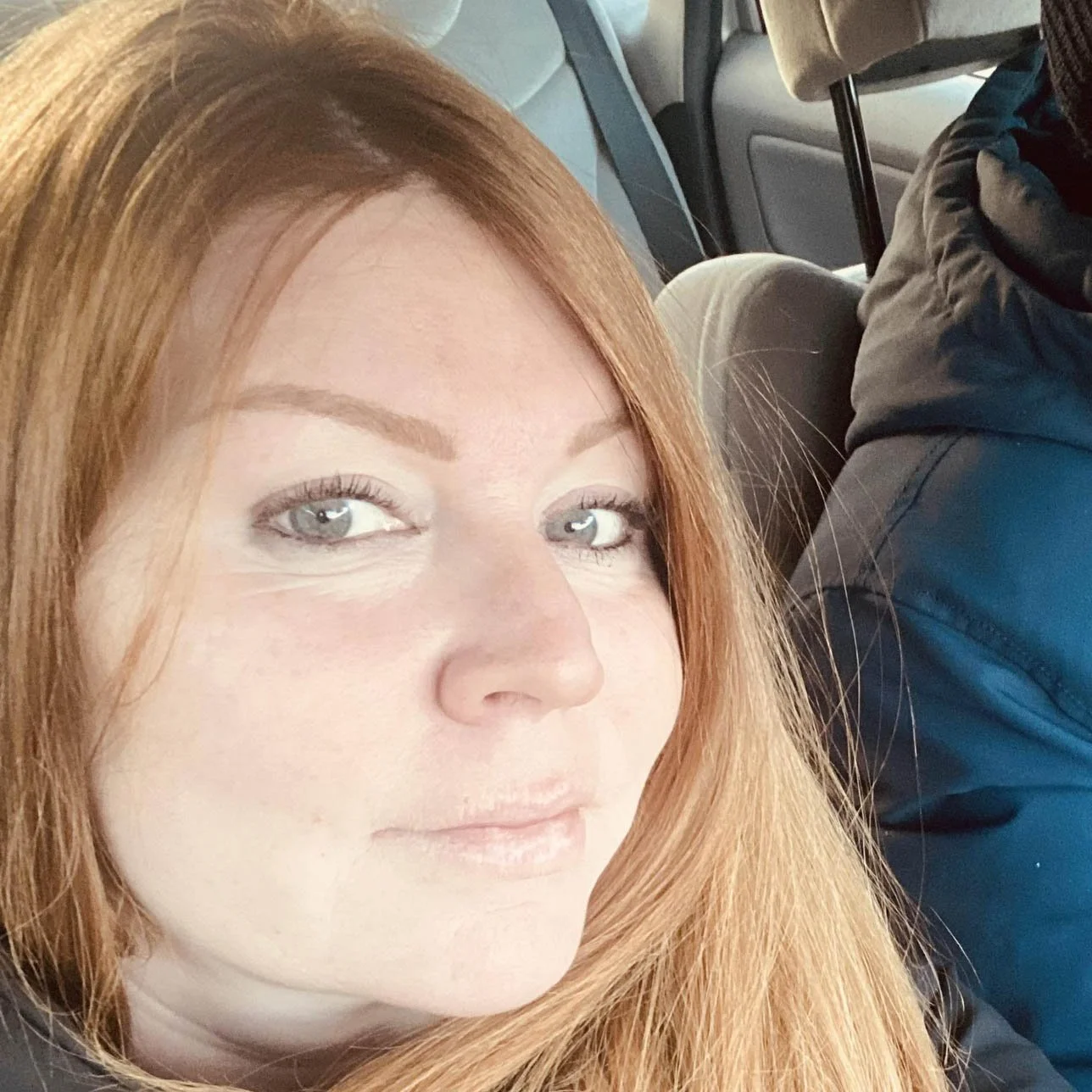 Une femme aux cheveux roux avec un regard bleu clair dans un véhicule, près d'une personne portant une veste noire.