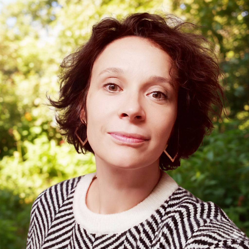 Une femme avec des cheveux courts et bouclés pose devant un fond de nature verdoyante.
